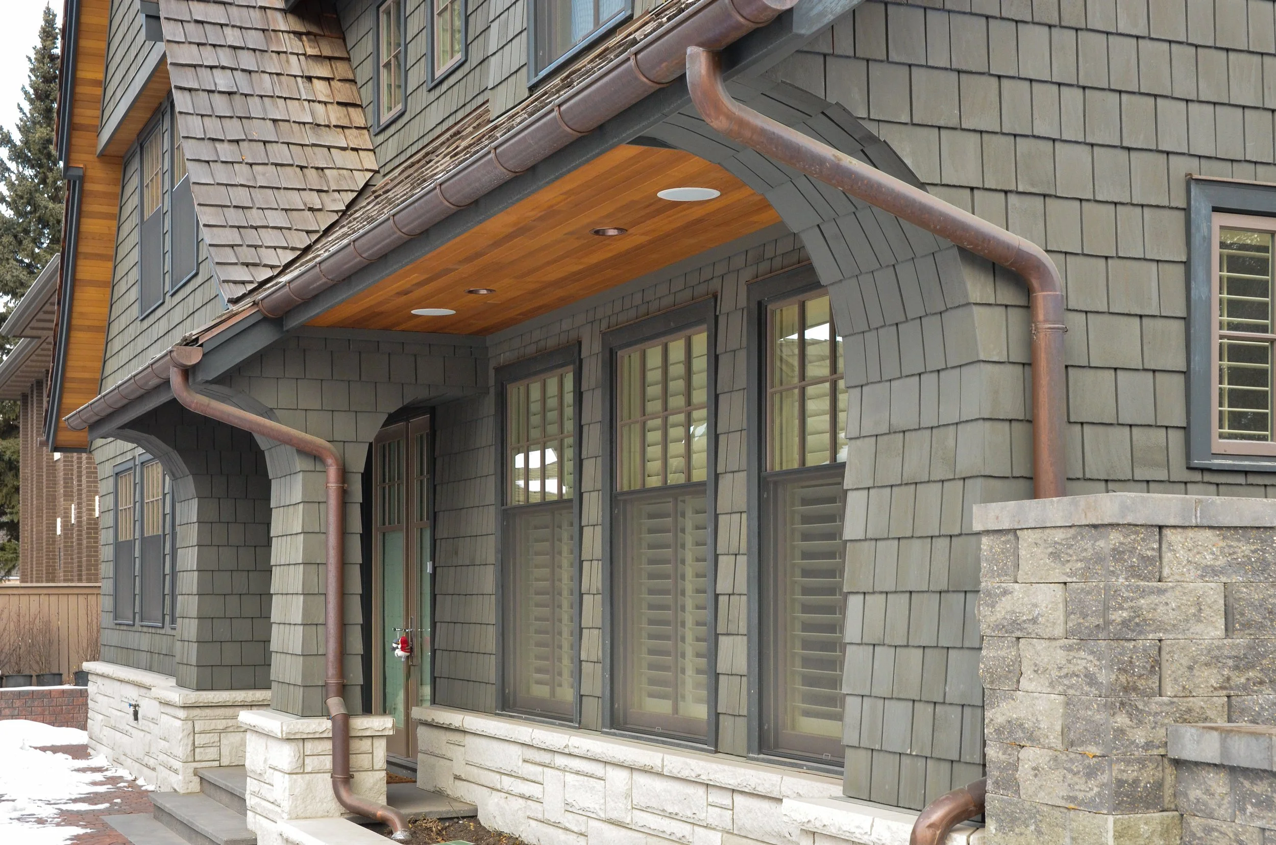 Close-up of a house exterior with green siding, large windows with shutters, stone porch, copper gutters, and a wooden ceiling on the porch