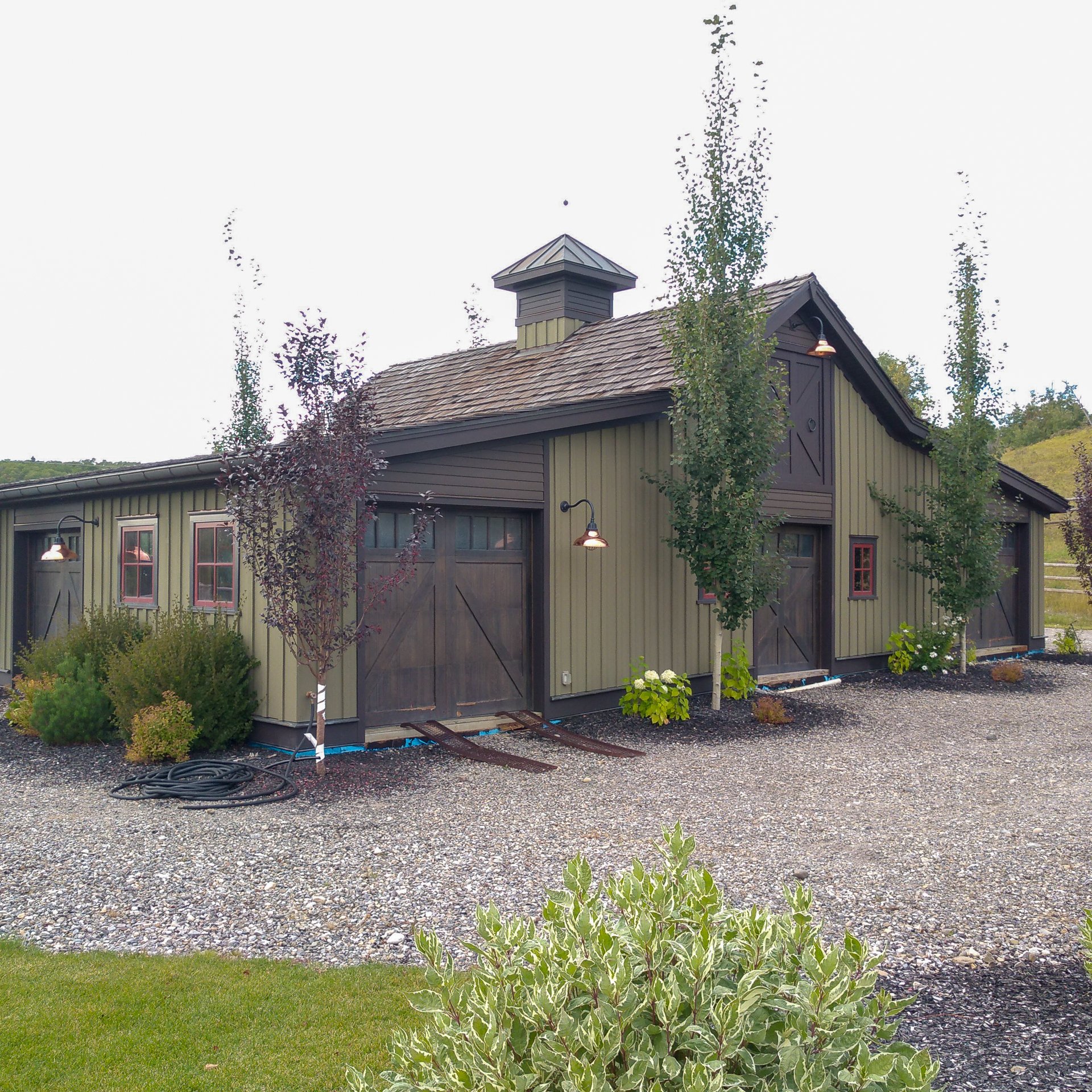 A rustic-style barn with green siding and dark brown doors, surrounded by young trees and shrubs, on a gravel yard with a small patch of grass in the foreground.