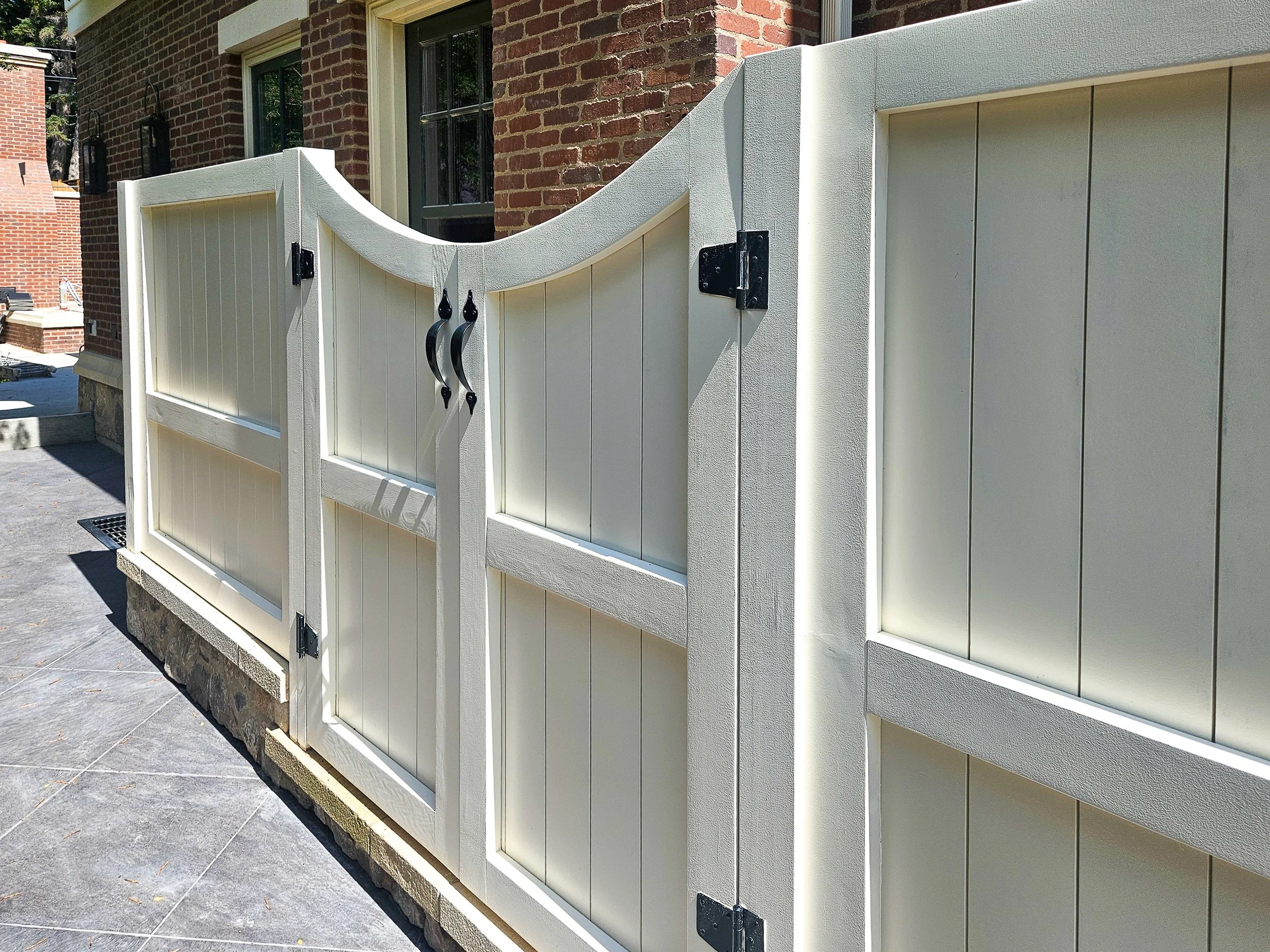 White wooden fence with decorative black metal hardware, attached to a brick house with two windows.