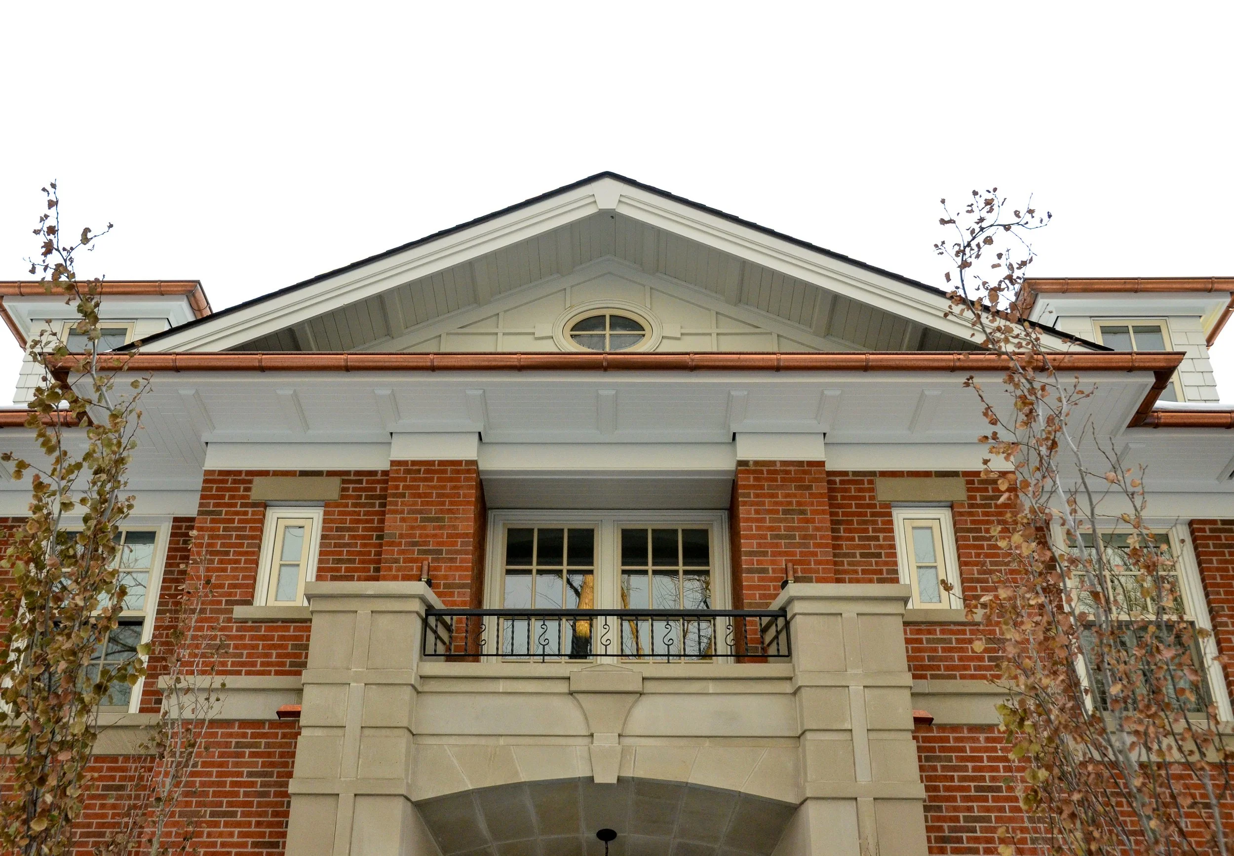 Front view of a large, brick and stucco house with a balcony and gabled roof, flanked by trees with leaves.