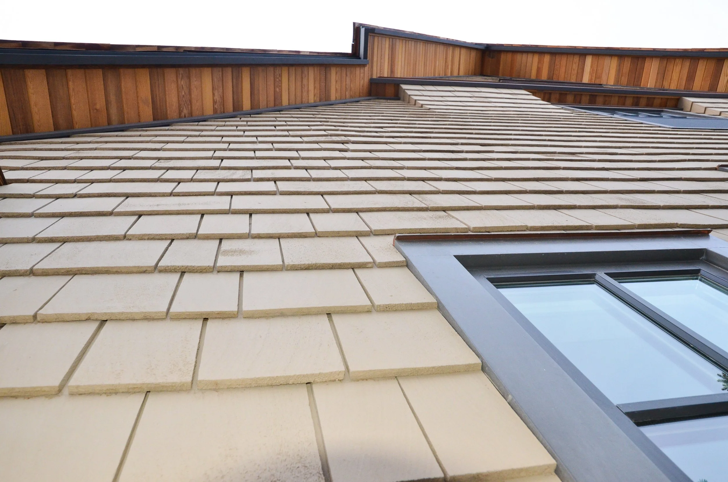 Close-up view of a house exterior showing beige brick siding, a window with gray trim, and wood paneling at the top under a cloudy sky.