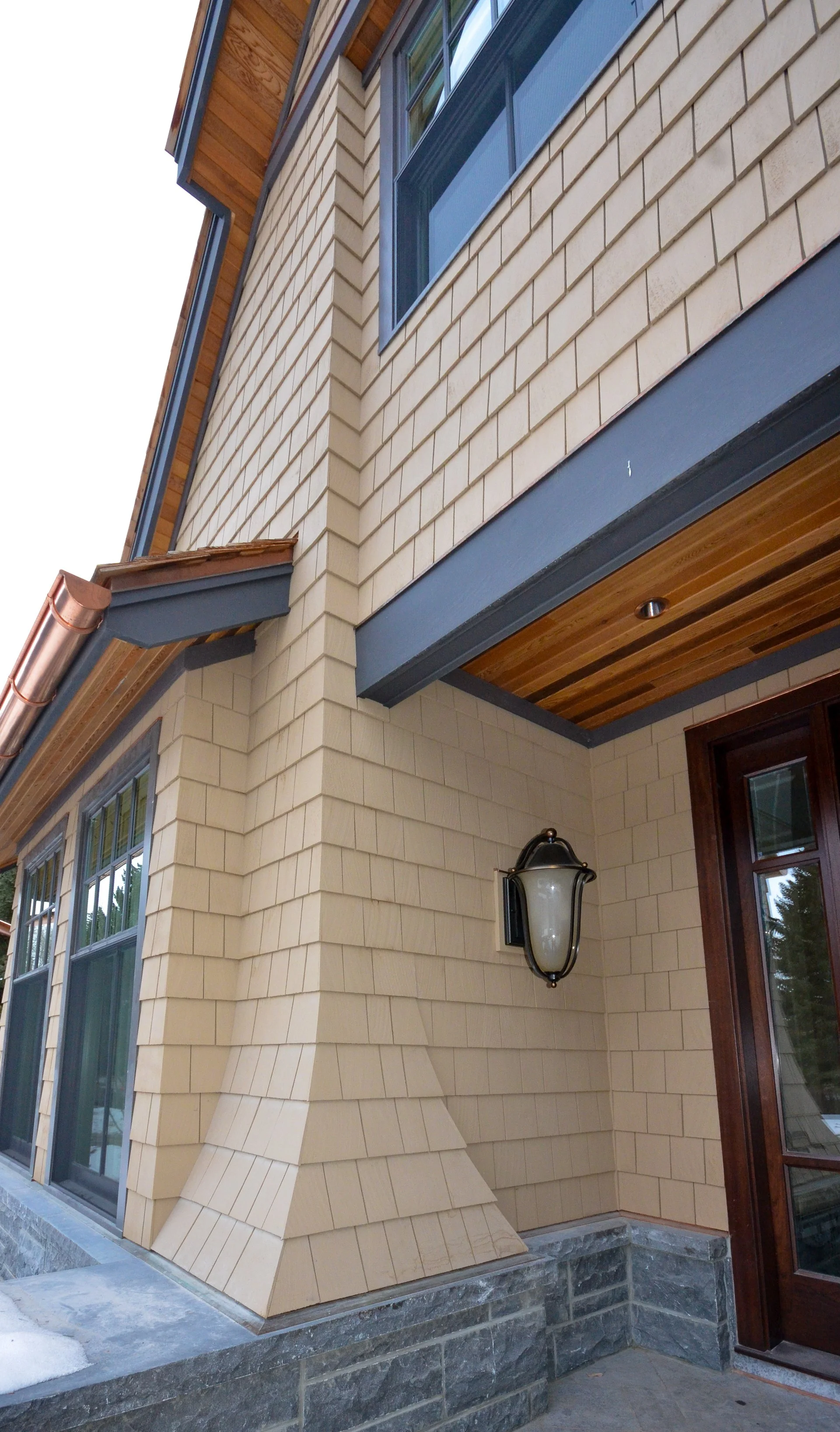 Close-up of a building's exterior showing beige siding, grey trim, a wooden ceiling, a wall-mounted outdoor lantern, and a stone foundation.