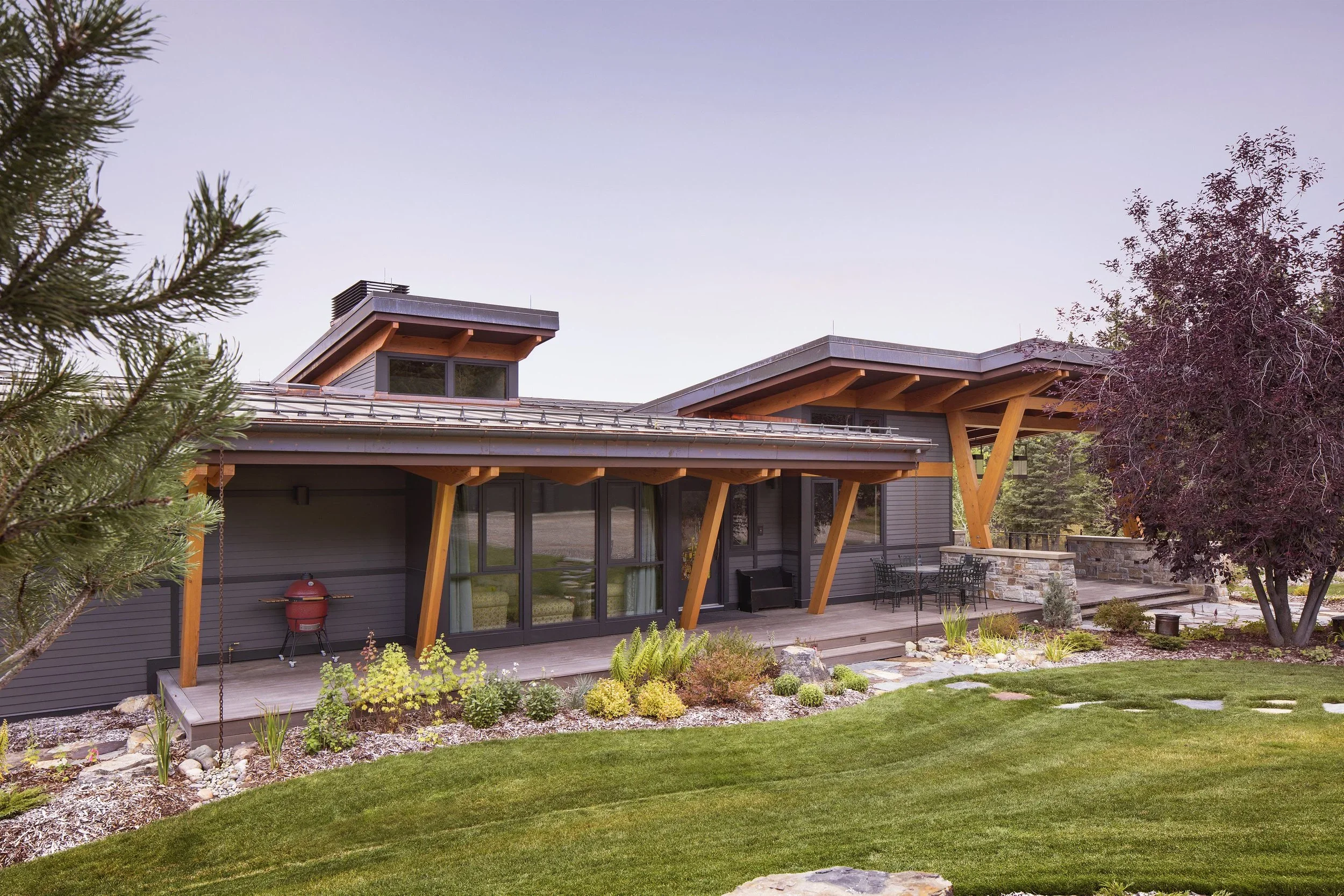 Modern house with a landscaped yard, featuring a patio with outdoor furniture and a red grill, surrounded by trees and greenery. Dda Architecture Ltd.