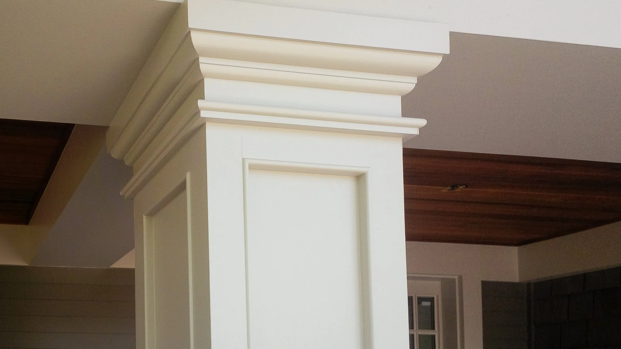 Close-up of a white decorative column with crown molding, set in an interior space with a wood ceiling and a window in the background.