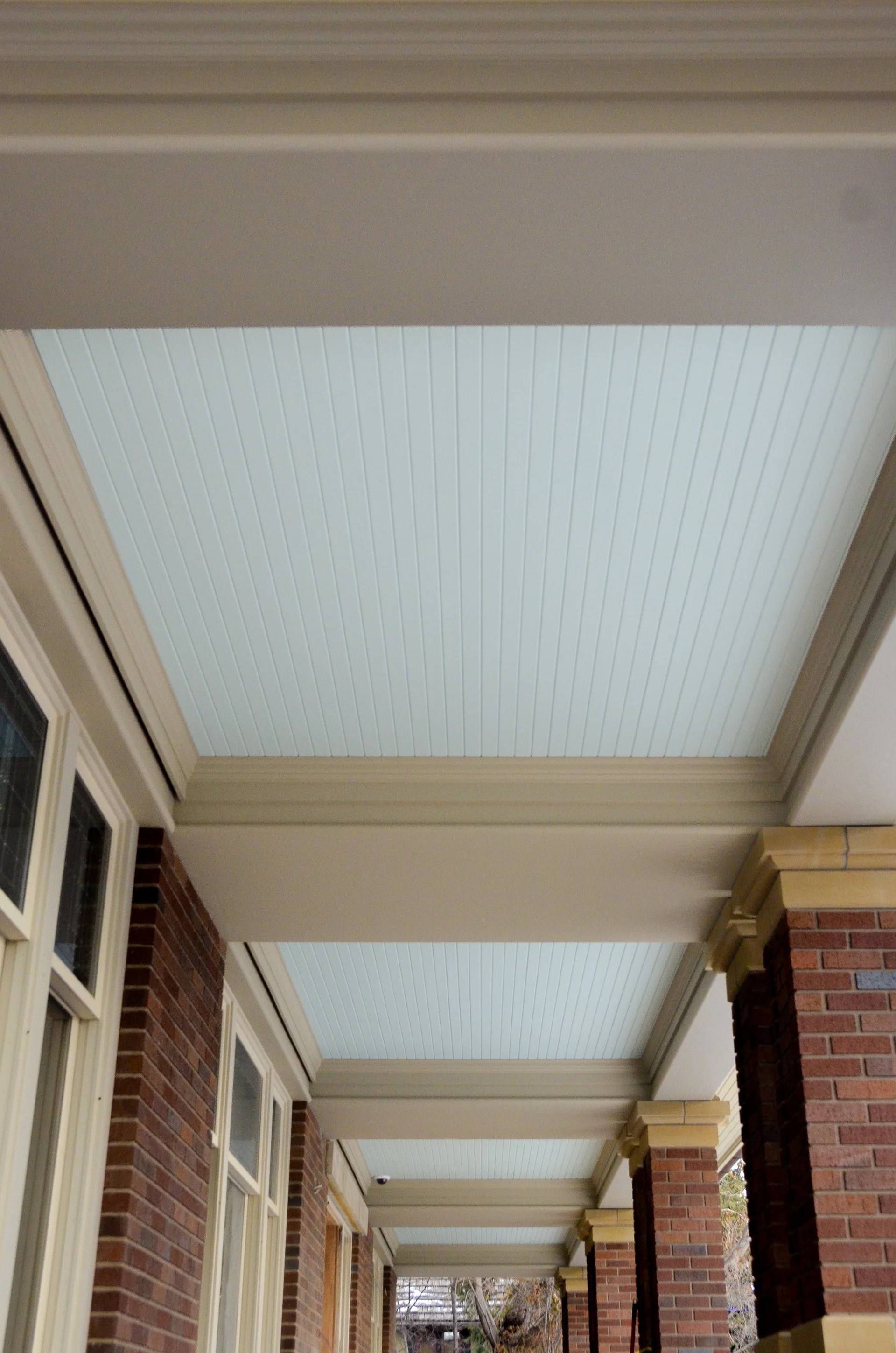 A porch ceiling with beige beams and white beadboard panels surrounded by brick columns and walls.