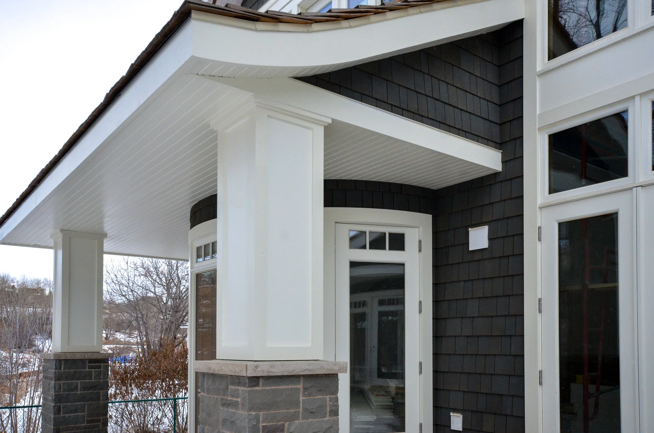 Close-up of a modern house exterior with dark shingles and white accents, including a door, windows, and tram siding, with trees and snowy landscape in the background.