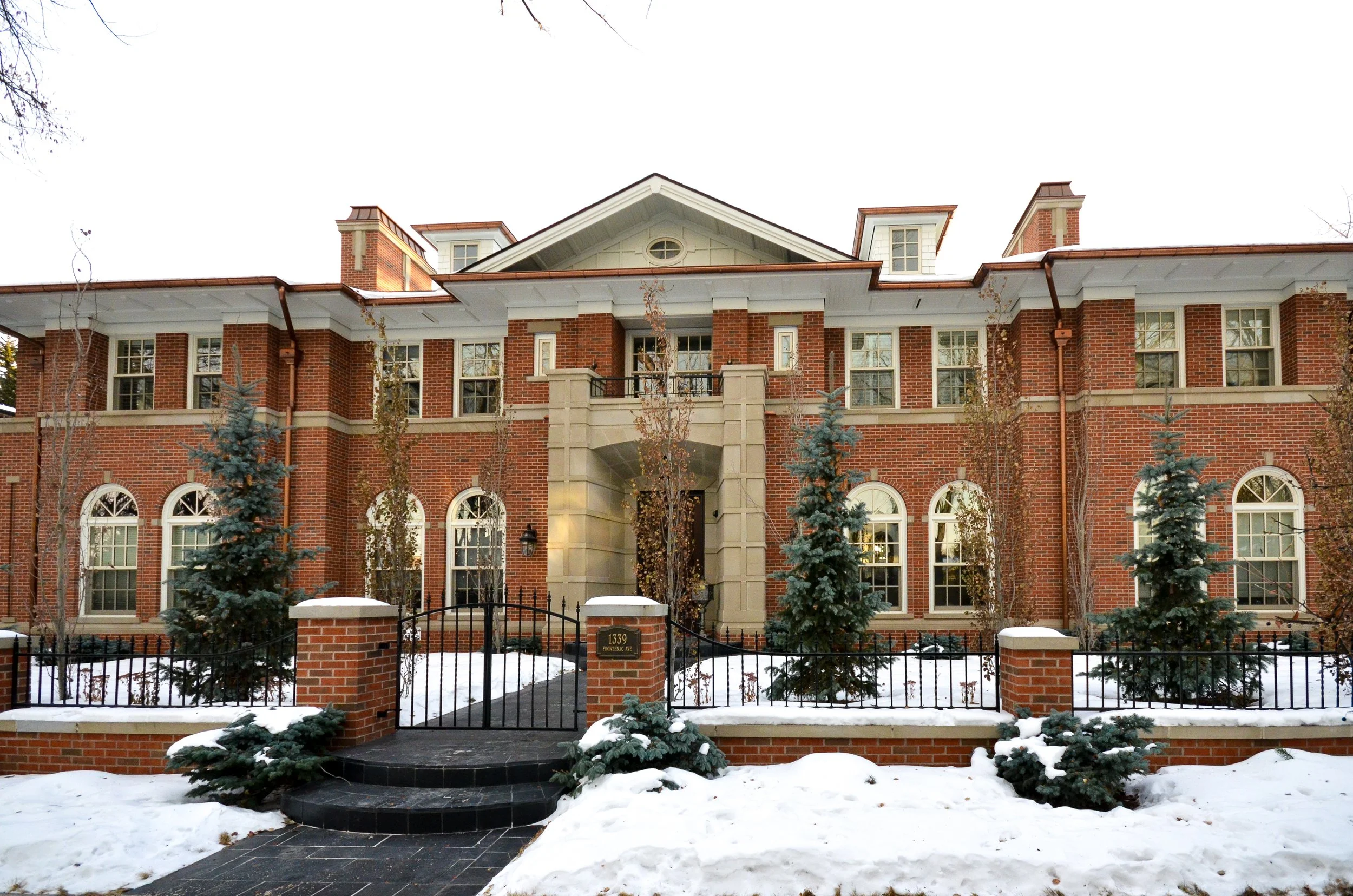 Front view of a large brick house with snow-covered ground and evergreen trees in winter.