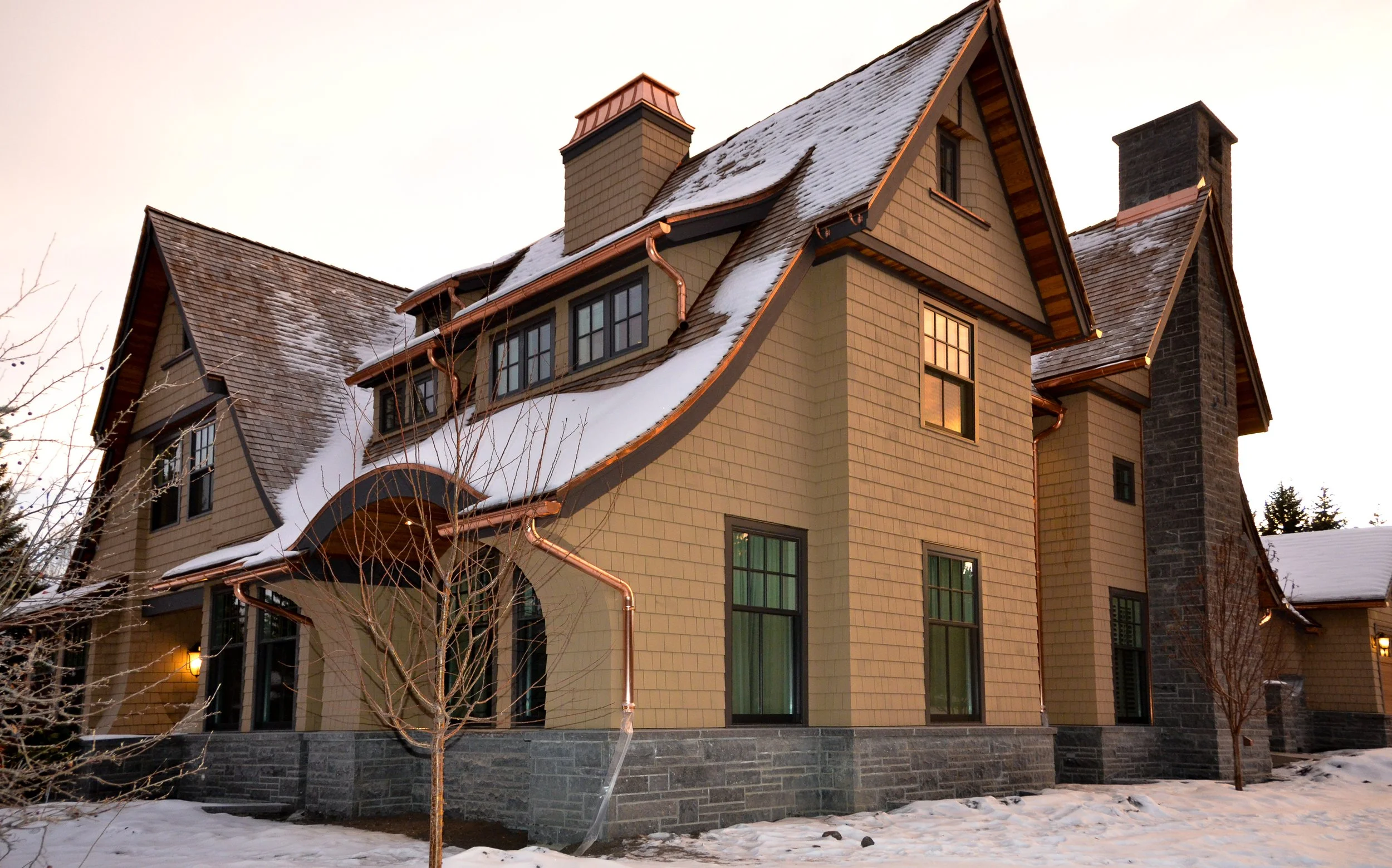 A large, beige, multi-story house with a steep roof, snow on the roof, and several large windows. The house has a stone foundation and a chimney, with leafless trees in the front yard and snow on the ground. Dda Architecture Ltd.