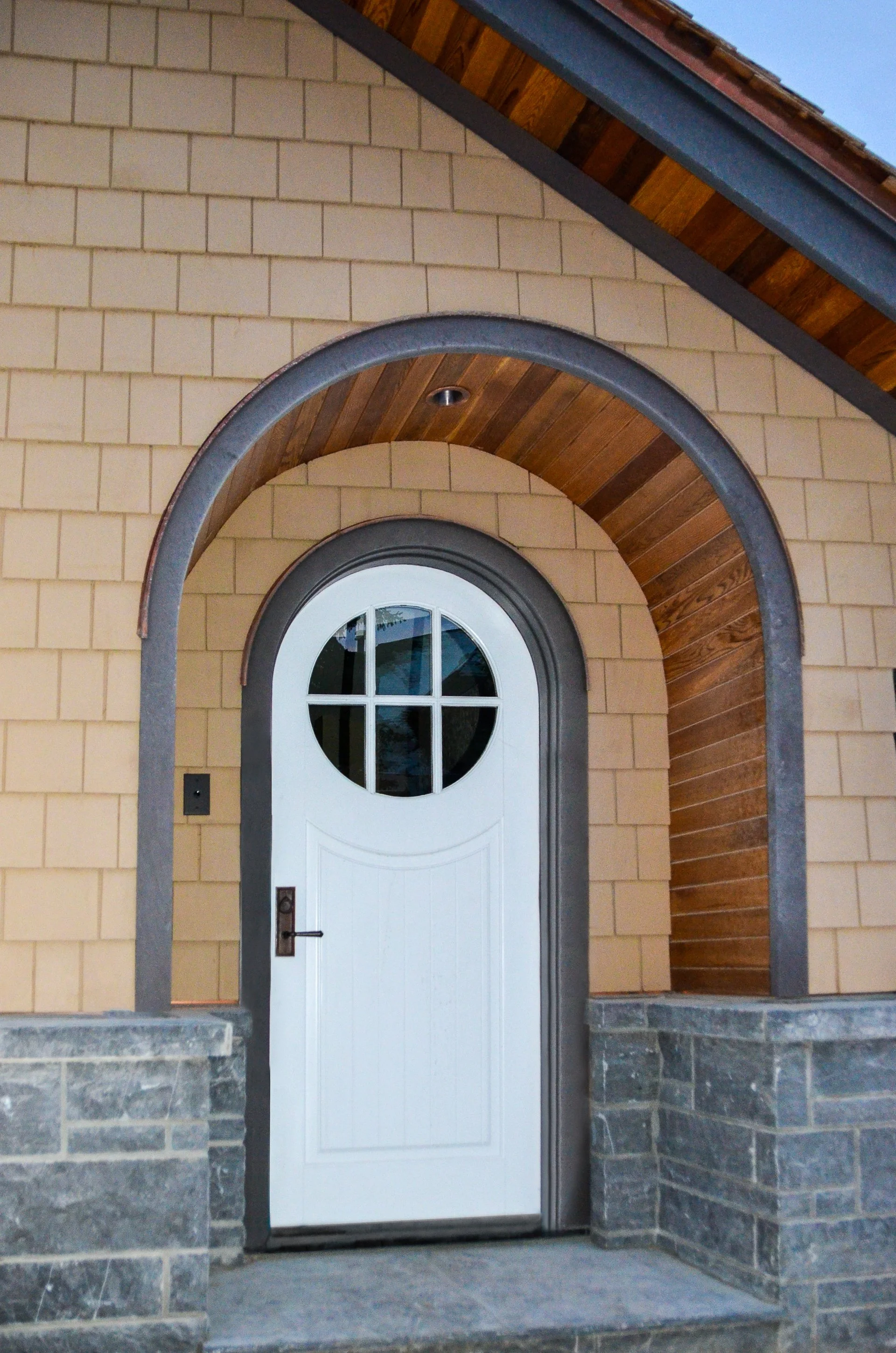 Front entrance of a house featuring a white door with a circular window and divided glass panes, surrounded by a stone and brick wall with wooden and metal accents. Dda Architecture Ltd.
