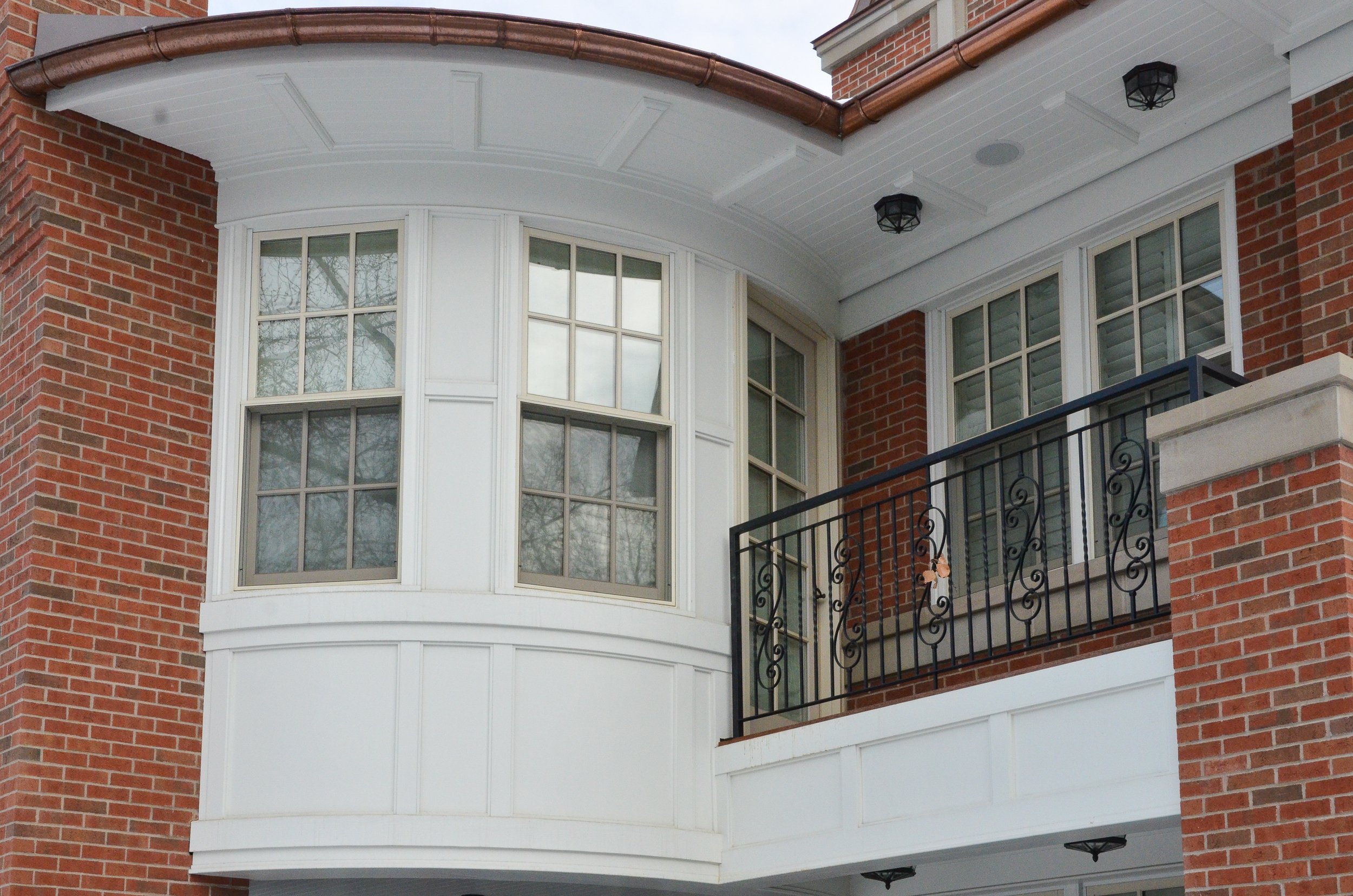 Close-up of a residential building's second-story corner with large windows, a small balcony with a decorative iron railing, and red brick facade with white trim under an overcast sky.