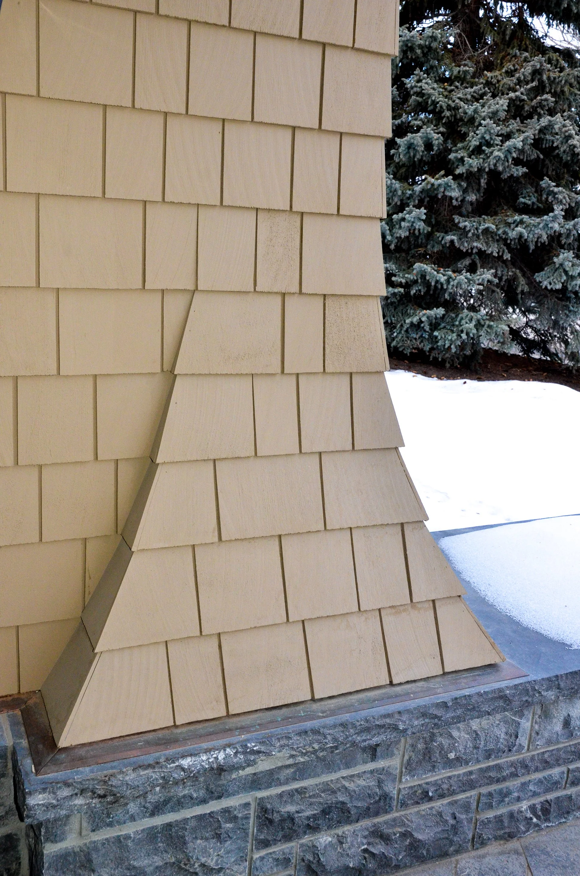 Close-up of a house chimney with beige shingles and a stone base, with snow and a pine tree in the background.