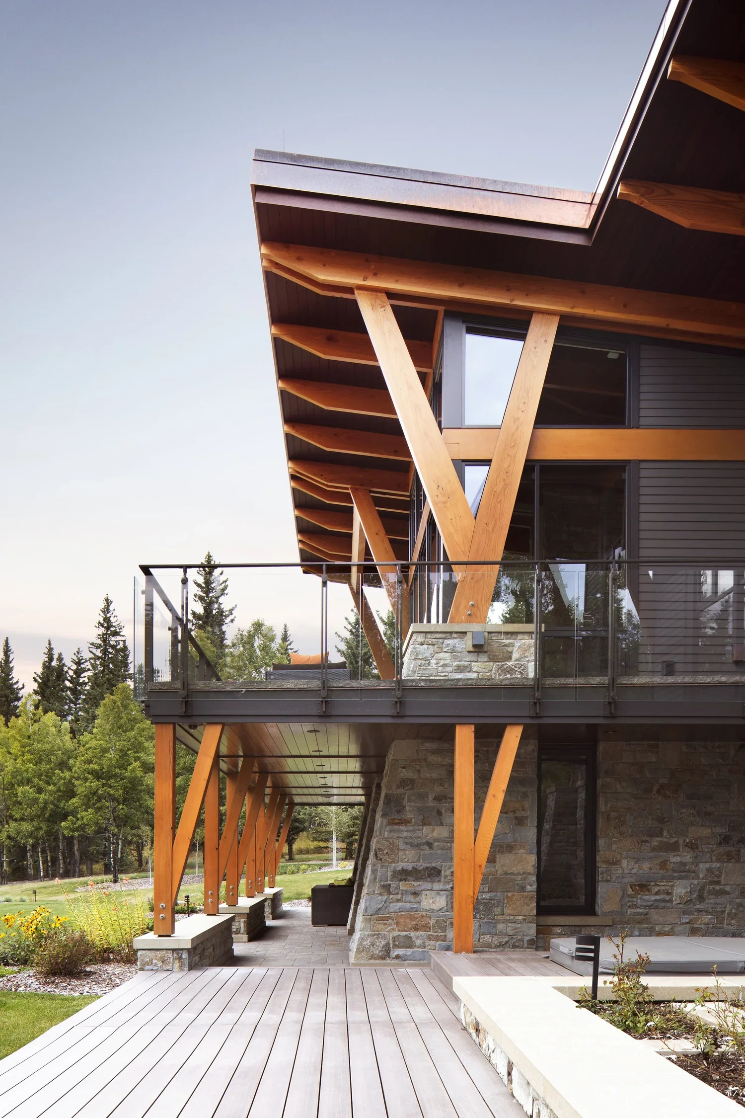 Modern multi-story house with wooden beams, glass balcony, stone walls, and large windows, situated in a lush green landscape. Dda Architecture Ltd.