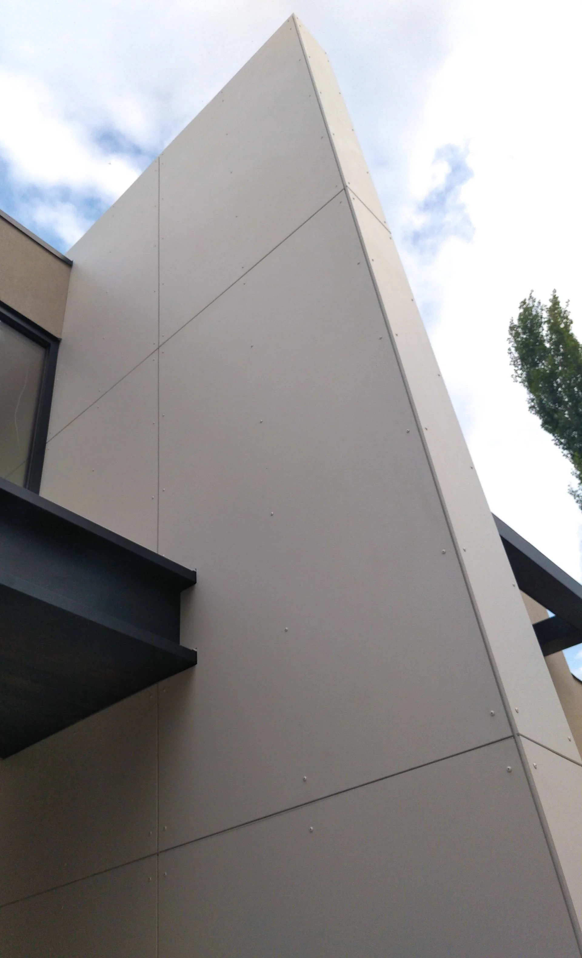 Close-up view of a modern building's exterior wall made of large, beige panels with visible nails and a small overhang in black. Part of a window is visible on the left, and trees with green leaves and a partly cloudy sky are in the background.