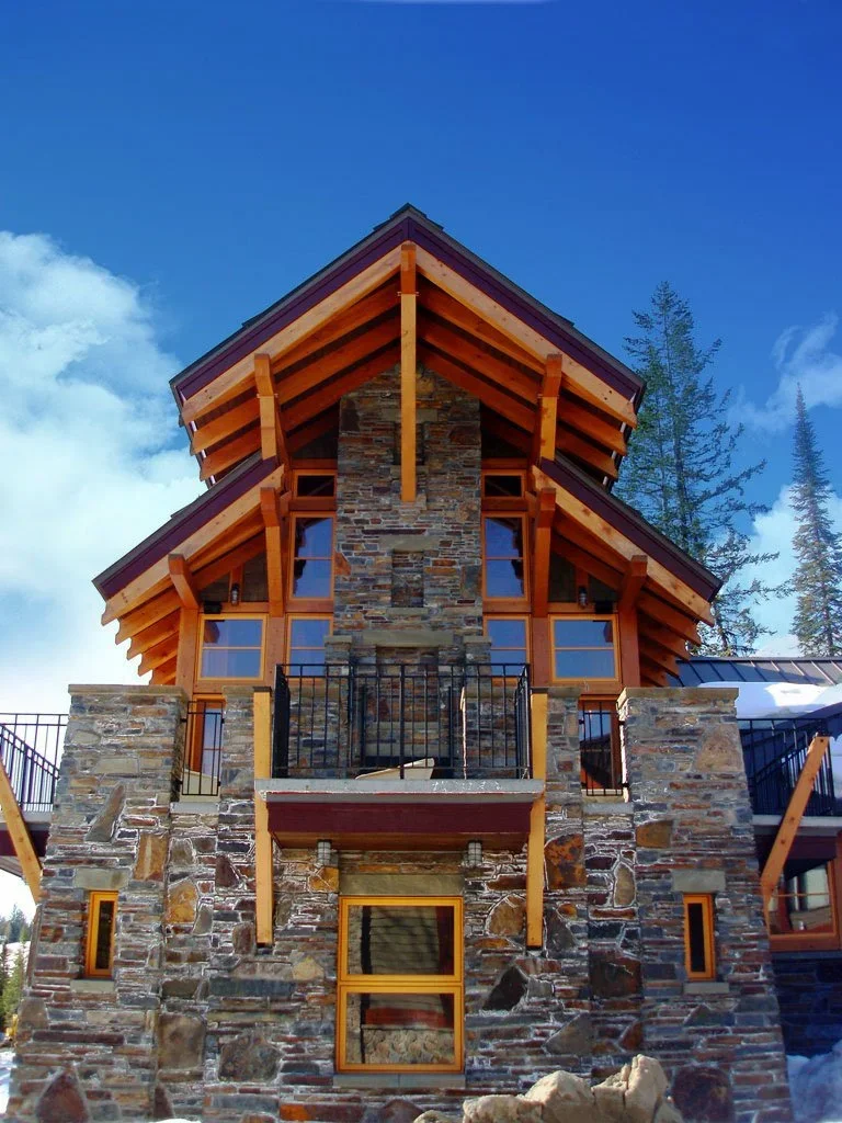 A mountain lodge-style house with stone and wood construction, multiple large windows, and a prominent central chimney, set against a bright blue sky and surrounded by tall trees. Dda Architecture Ltd.