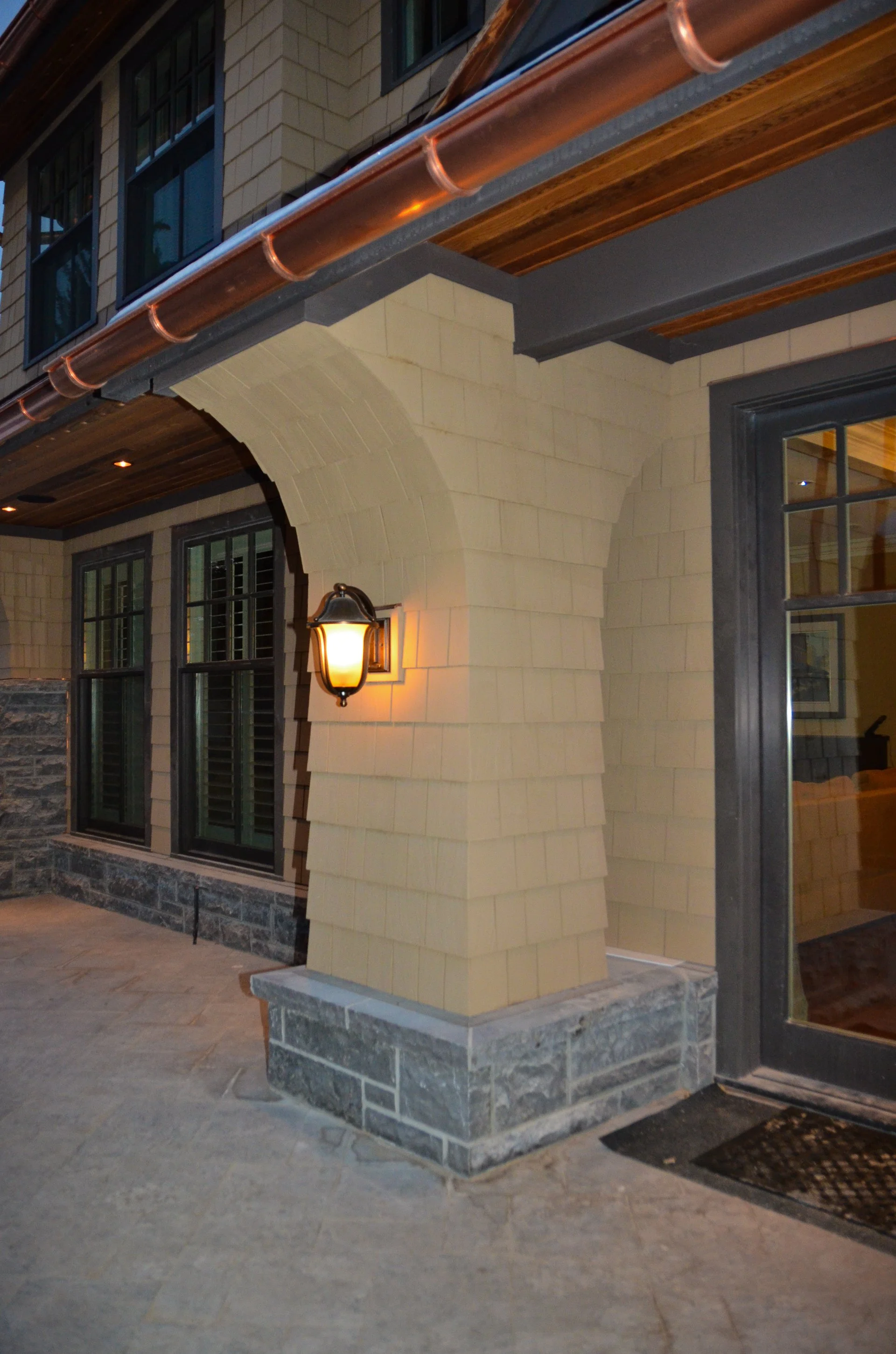 Close-up of a residential house's exterior corner at night, showing stone and siding, windows with shutters, a wall-mounted lantern light, and a sliding door with black framing.