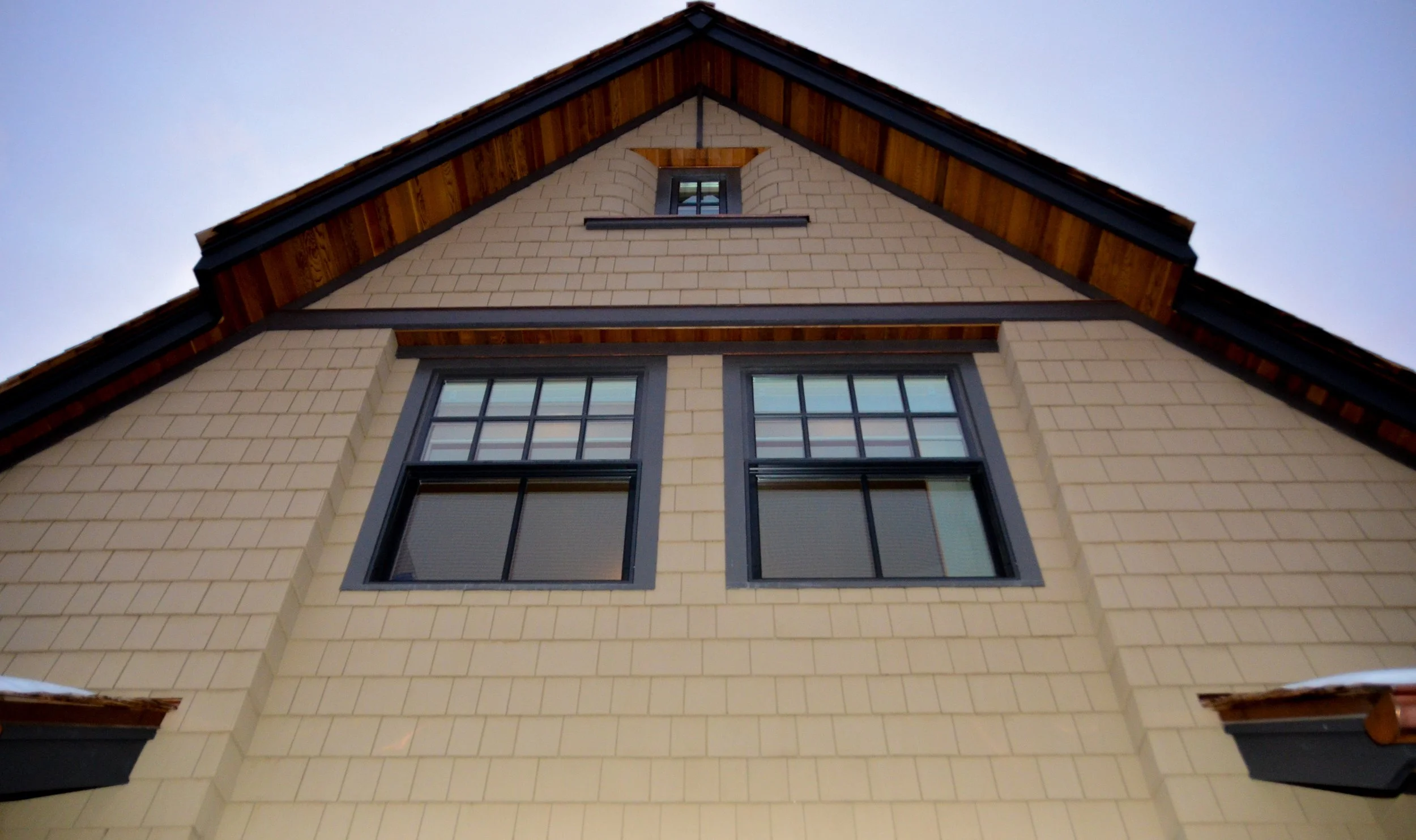 View from below of a house with beige brick exterior and dark window frames, a gabled roof with wooden accents, and small attic window. Dda Architecture Ltd.
