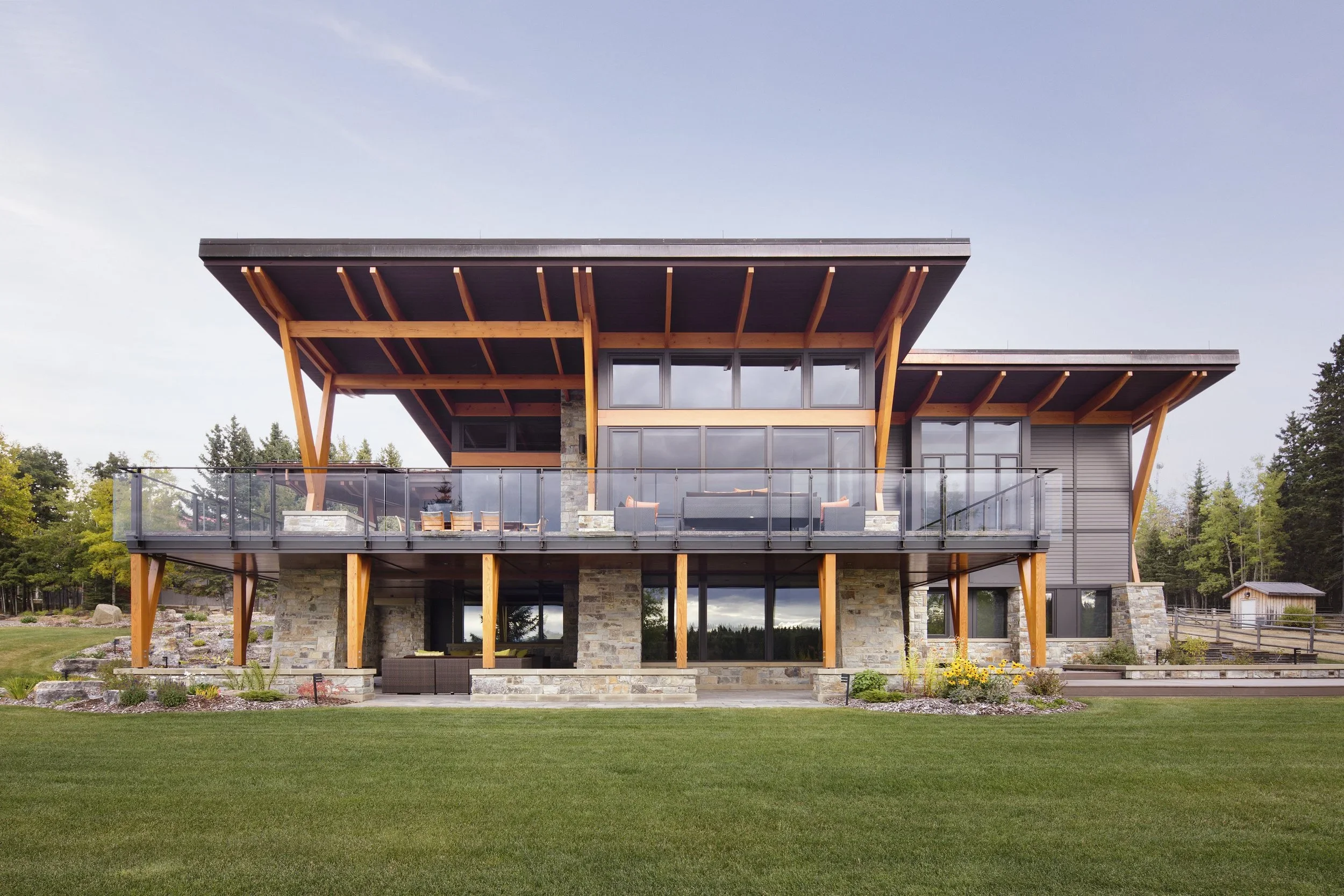 Modern, two-story house with large glass windows and a spacious balcony, featuring wooden beams and stone accents, surrounded by a well-manicured lawn and trees in the background. Dda Architecture Ltd.