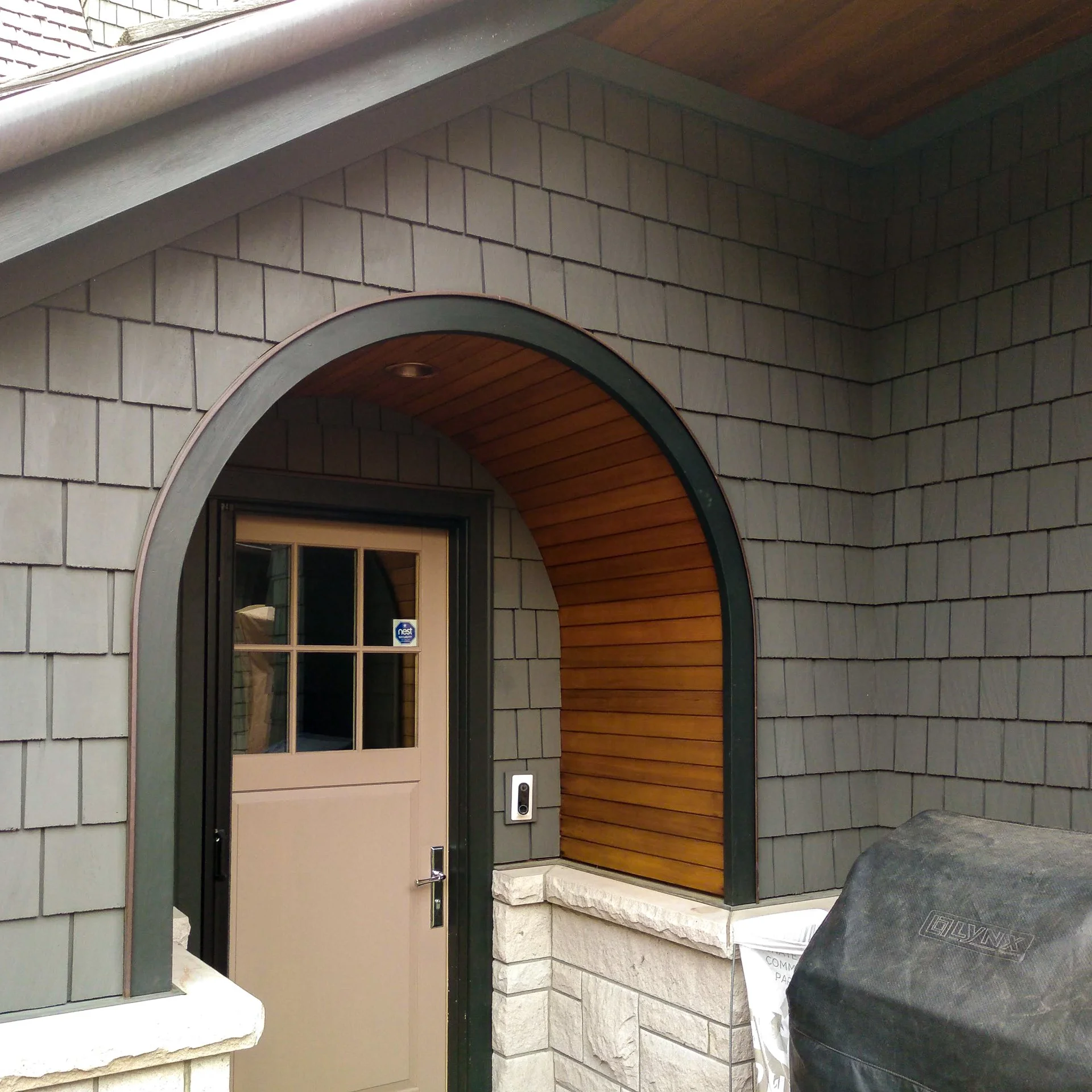 Exterior of a house with a beige door, arched wood-paneled alcove, gray shingle siding, and a stone facade. A black security camera is mounted near the door.