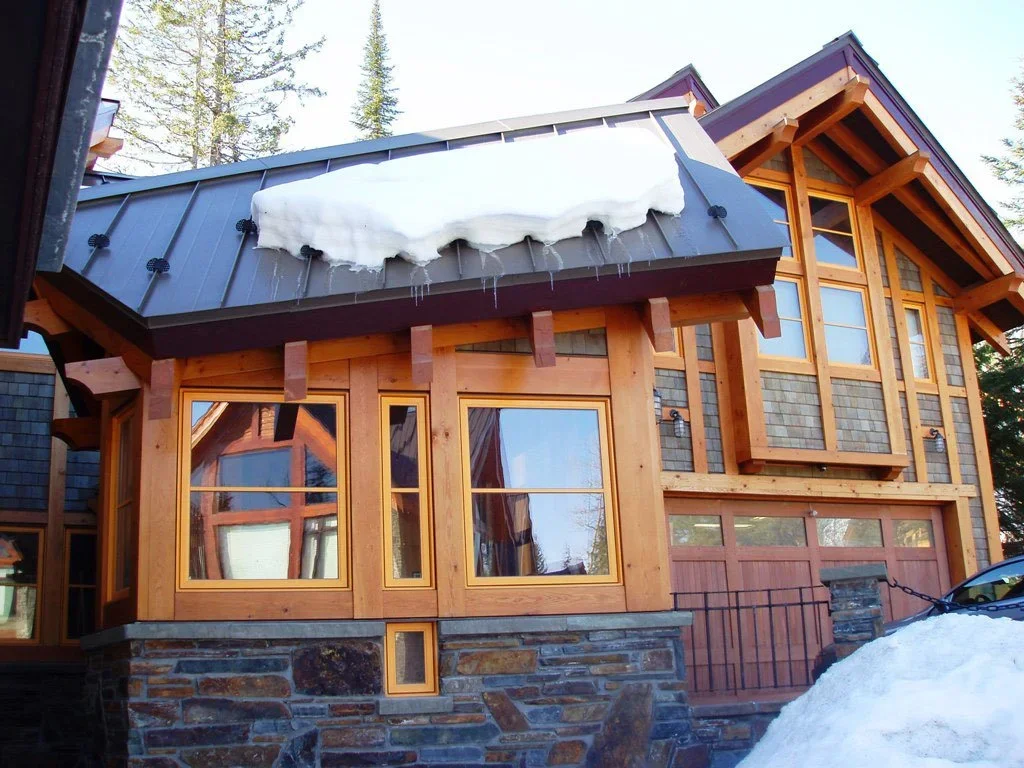 A wooden house with large windows, a stone foundation, and a metal roof partially covered with snow and icicles, surrounded by trees. Dda Architecture Ltd.