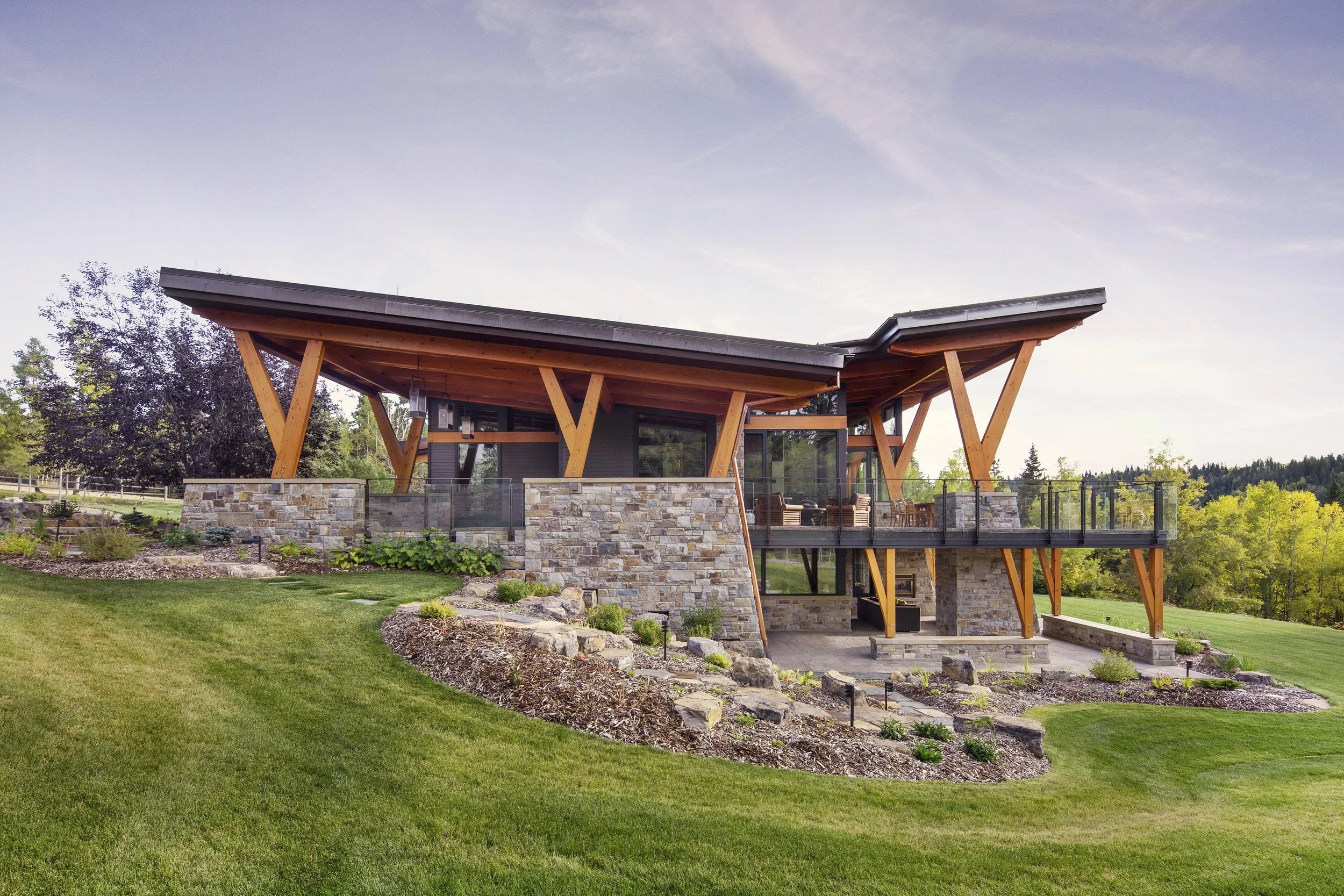 A modern house with large glass windows, wooden beams, and stone walls, surrounded by a landscaped yard with green grass and plants. Dda Architecture Ltd.