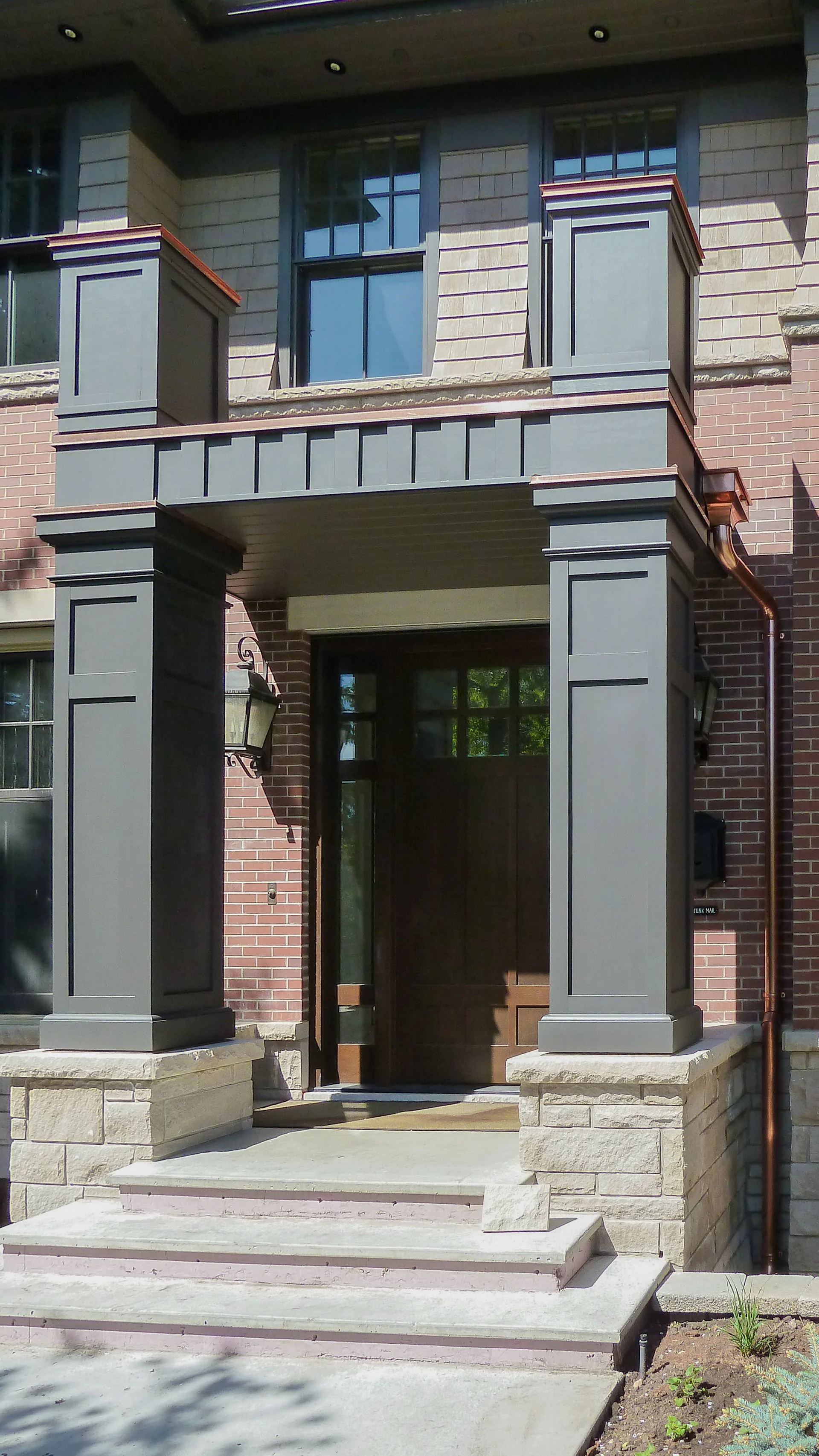 Residential house entrance with dark painted columns, stone steps, a wooden front door, and brick and siding exterior walls.