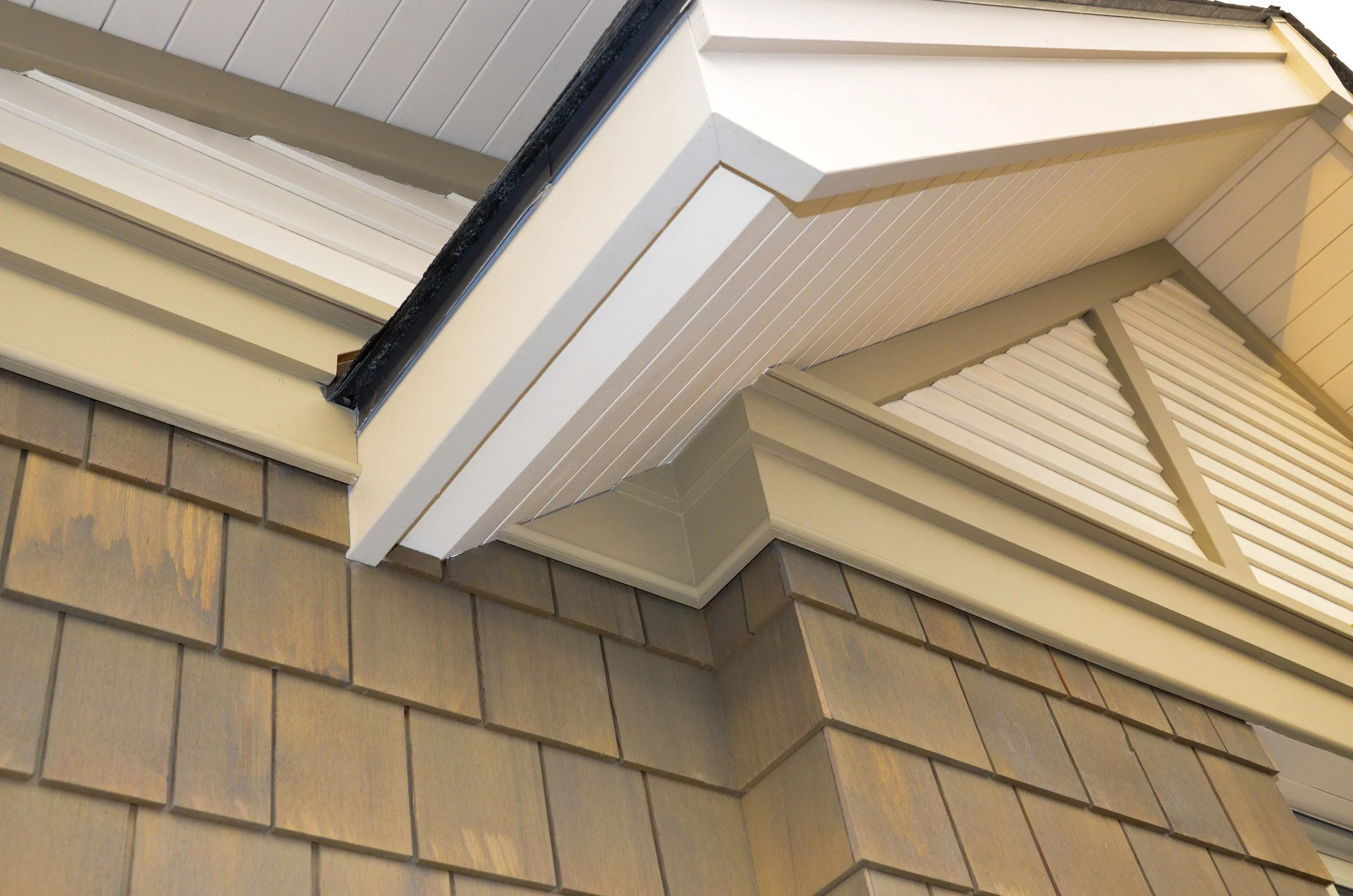 Close-up of the exterior corner of a house showing beige shingle siding, white trim, and venting around the roofline.