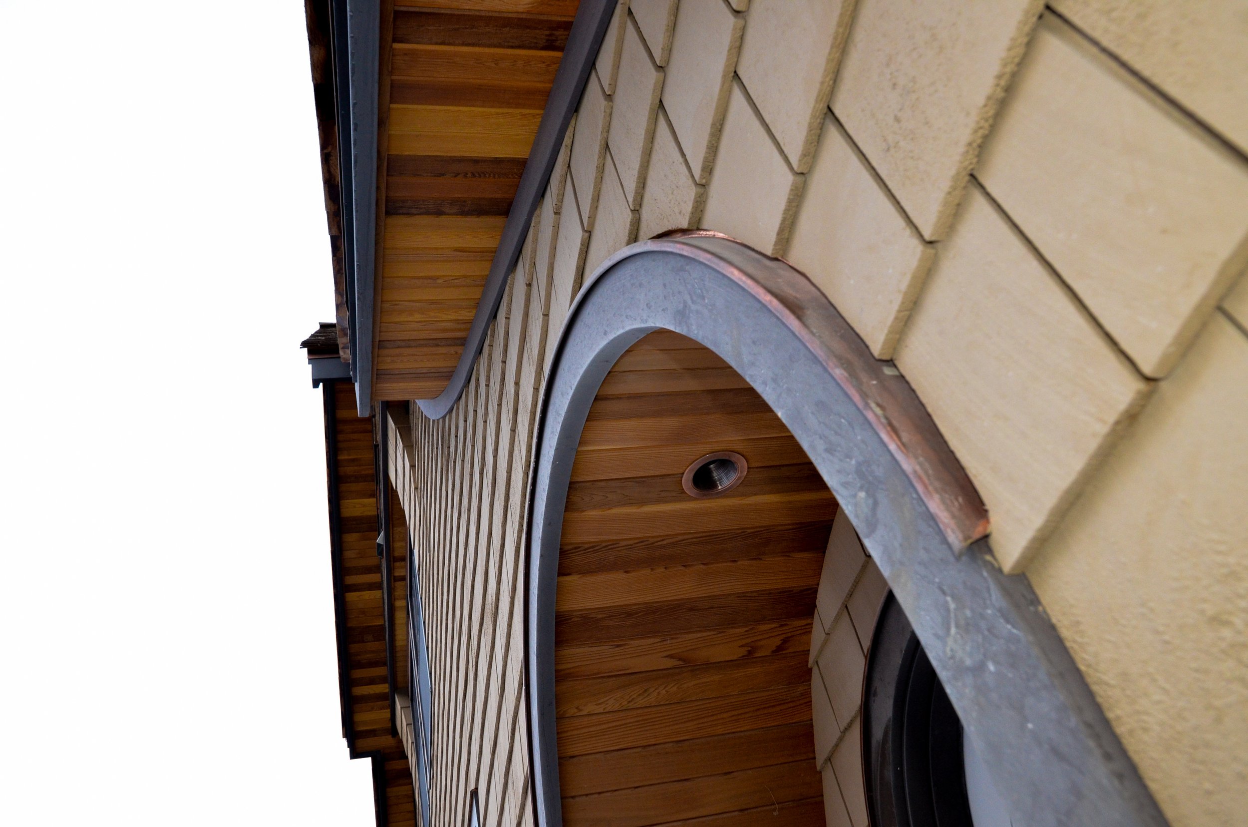 Close-up of a building exterior showing yellow brick wall, wooden siding, and a rounded metal window frame with copper trim, under a wooden roof with black edging.