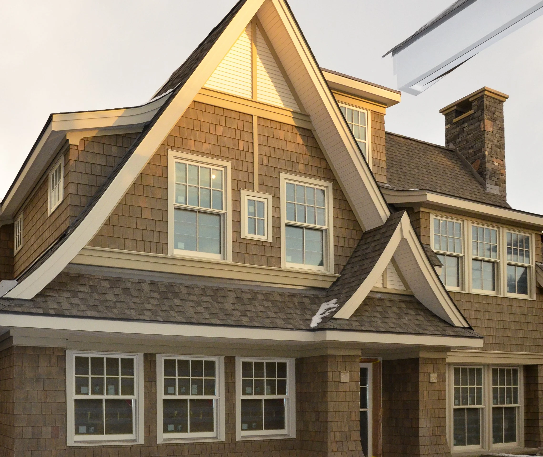 A large, multi-story house with brown shingle siding, multiple gabled roofs, and several white-framed windows. The house features a stone chimney and a small section of snow on the roof.