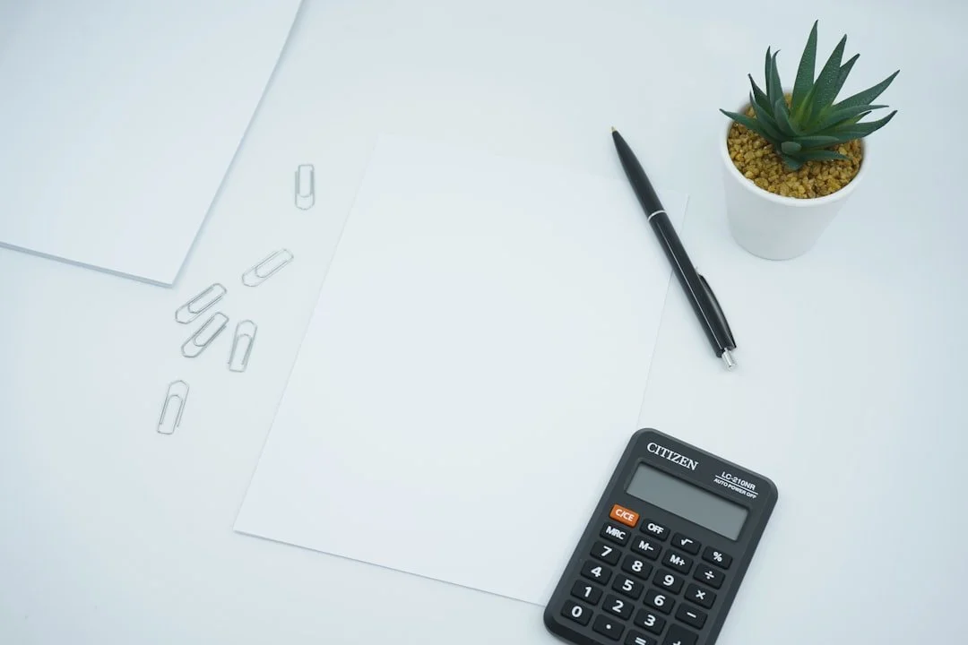A white desk with a few paper clips, a blank sheet of paper, a black pen, a calculator, and a small potted succulent plant.