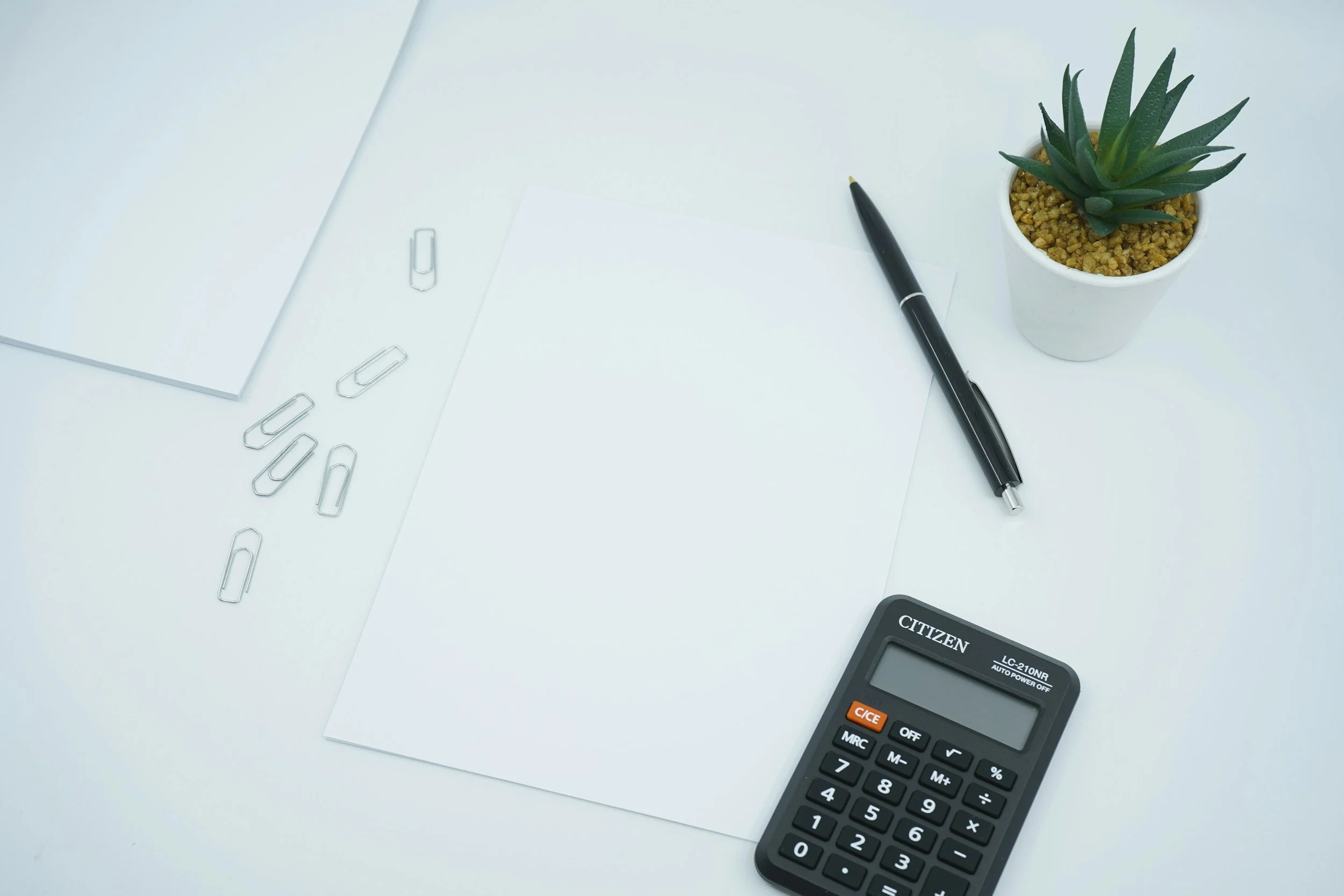 A white desk with a notebook, scattered paper clips, a black pen, a small potted cactus, and a black calculator.