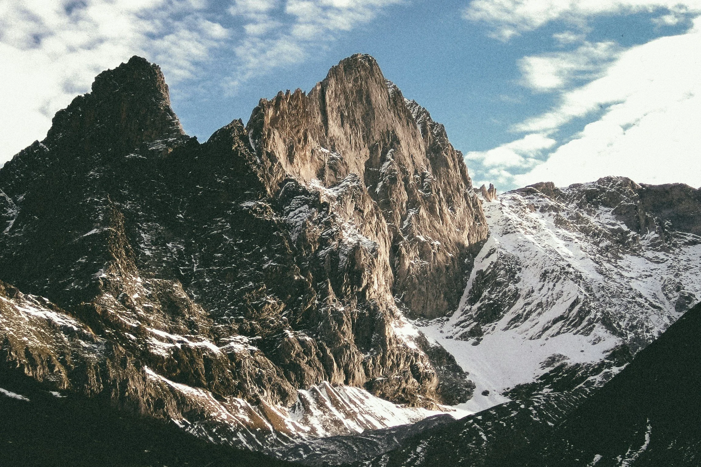 A rugged mountain with snow patches under a partly cloudy sky.