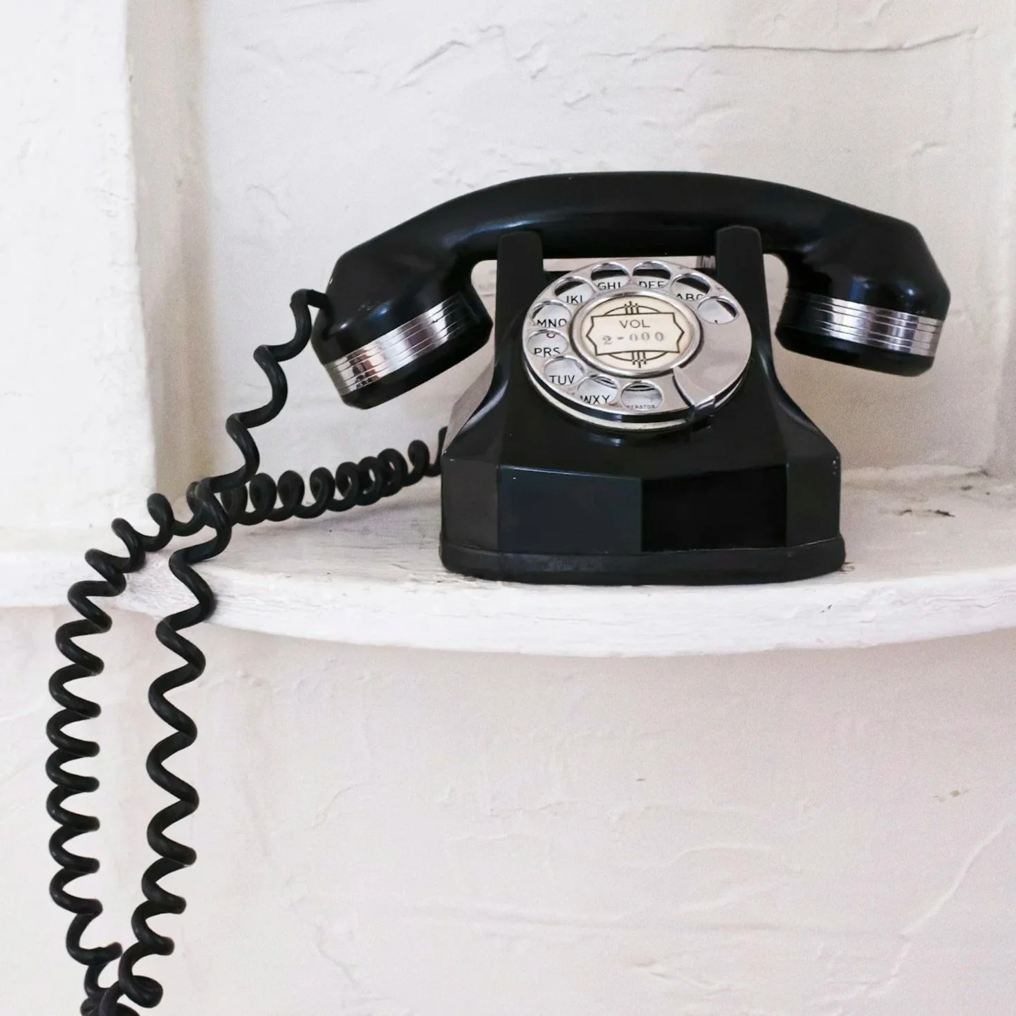 Vintage black rotary telephone with silver accents and coiled cord, placed on a white shelf.