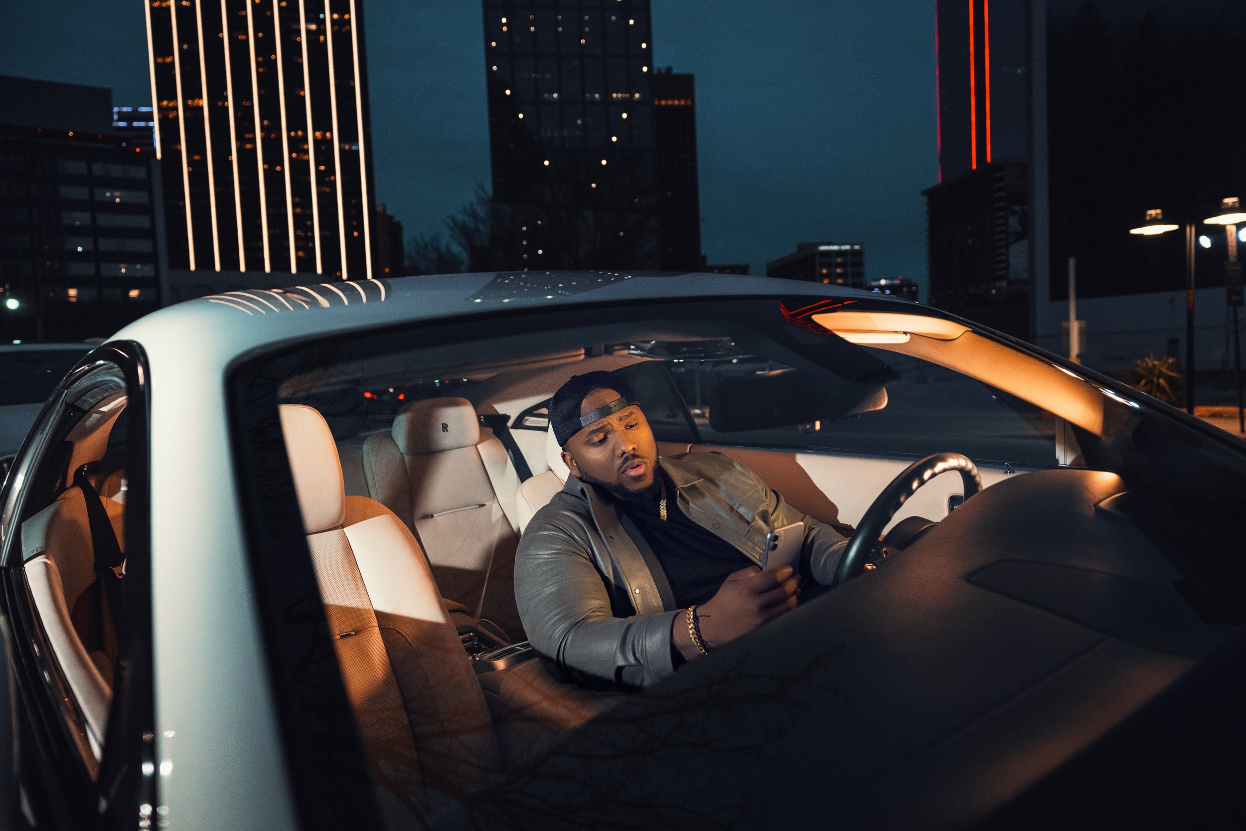 A man in a baseball cap and leather jacket sitting inside a car at night, looking at his phone with a city skyline in the background.