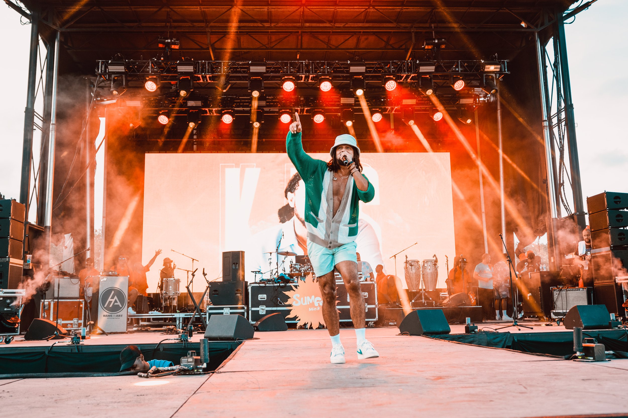 Performer singing on stage with a microphone, wearing a hat, green jacket, and shorts, during a concert with stage lights and backup musicians behind him.