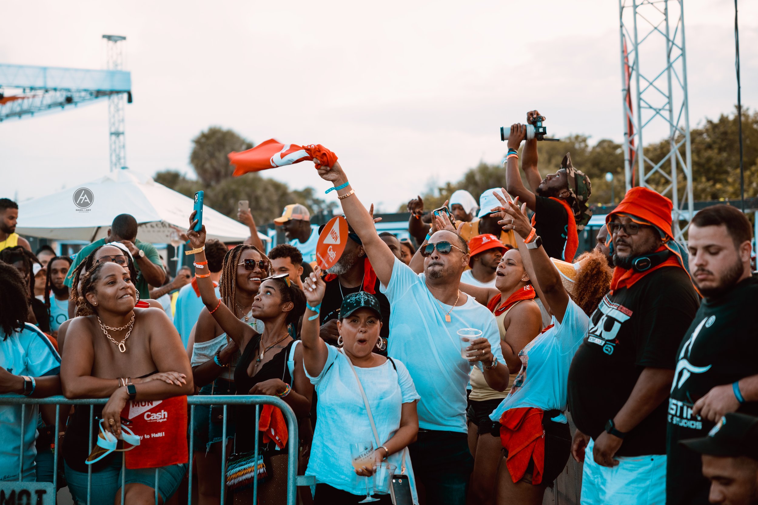 Crowd of diverse people at an outdoor event, some taking photos and videos, wearing casual summer clothing, with a person waving a red and white flag. There is a stage in the background and a fence separating the crowd.