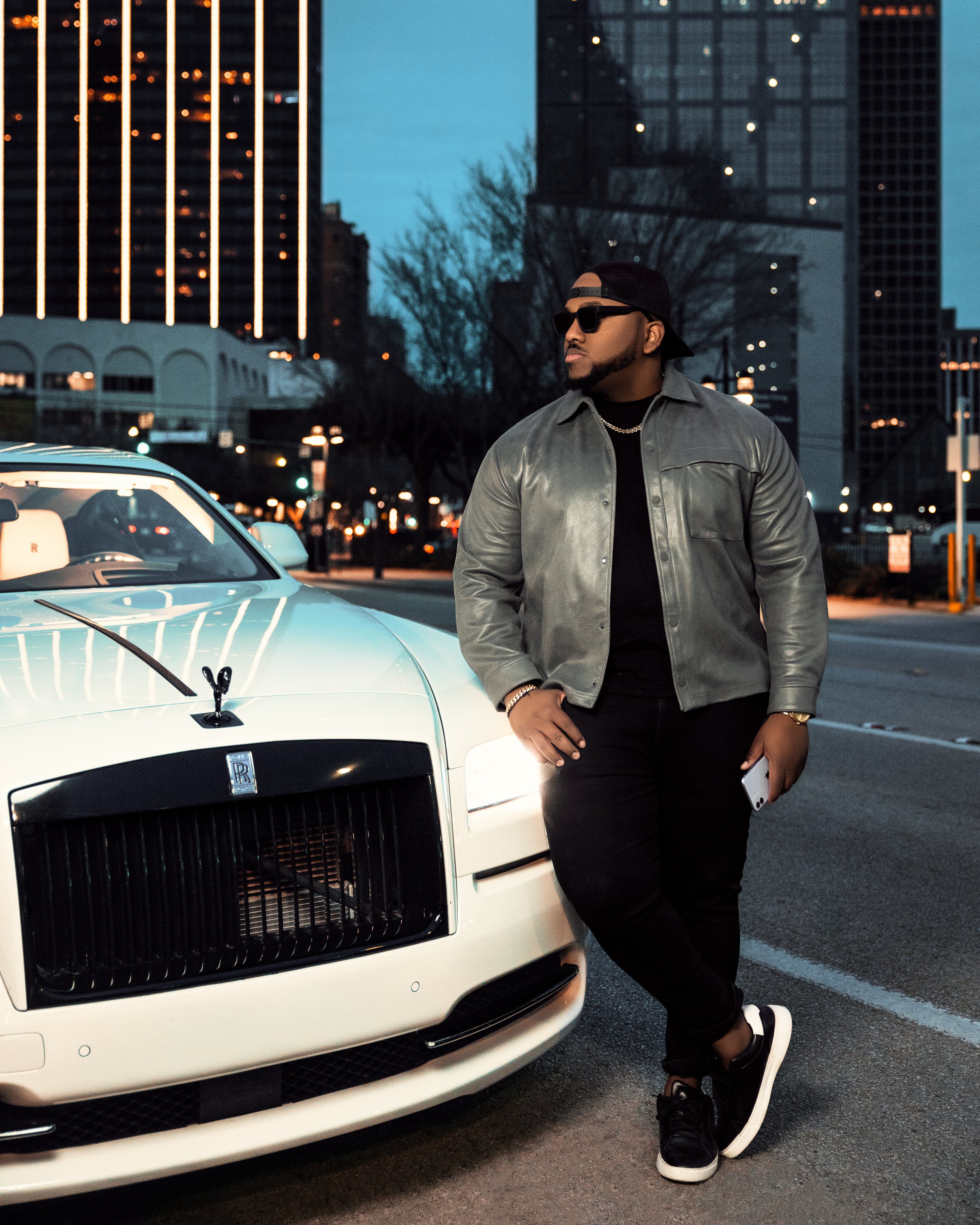 A man wearing a gray leather jacket, black pants, and black sneakers leaning against a white Rolls-Royce car on a city street during dusk, with tall buildings and city lights in the background.