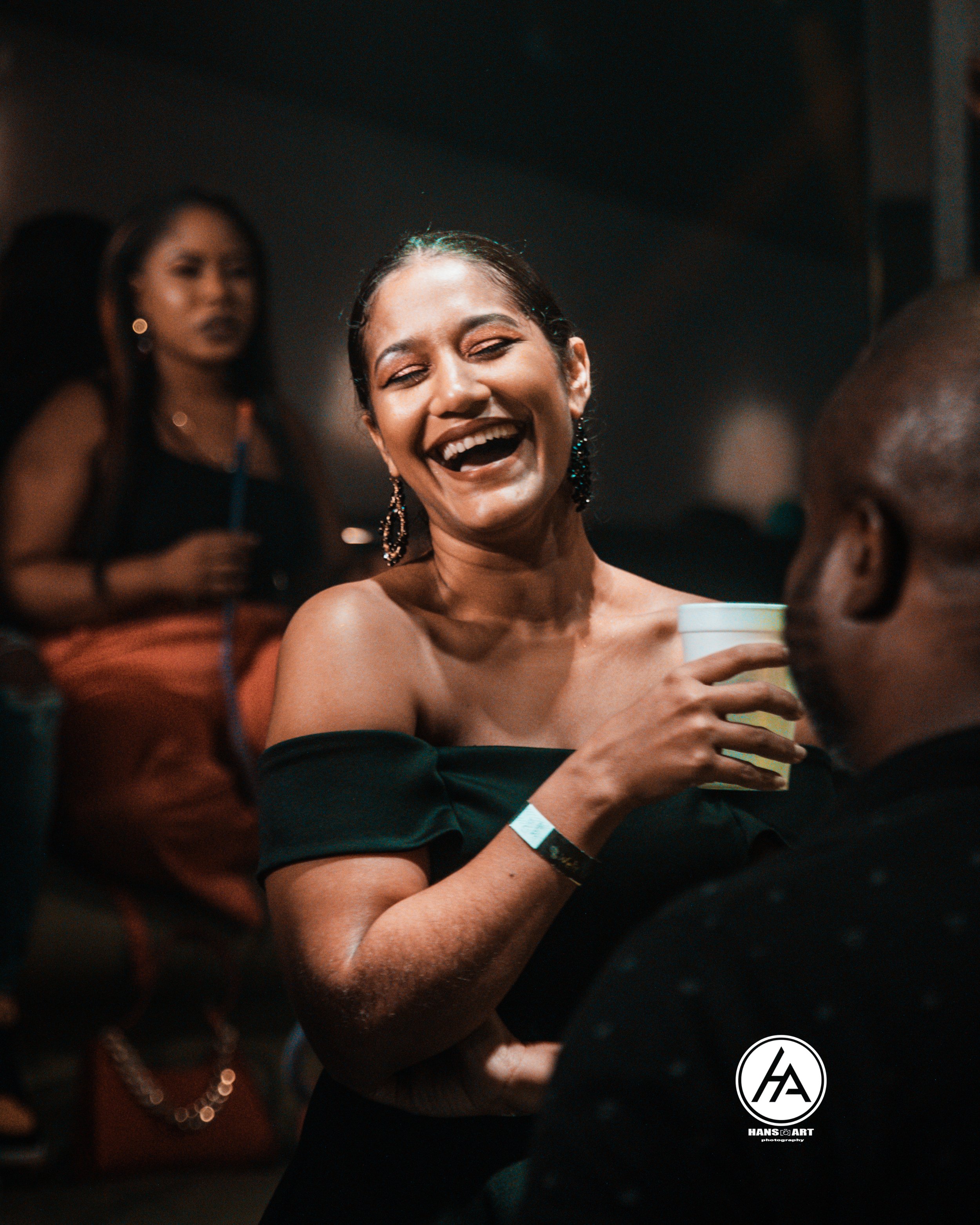 A woman with earrings smiling and holding a cup at a social event with other people in the background.