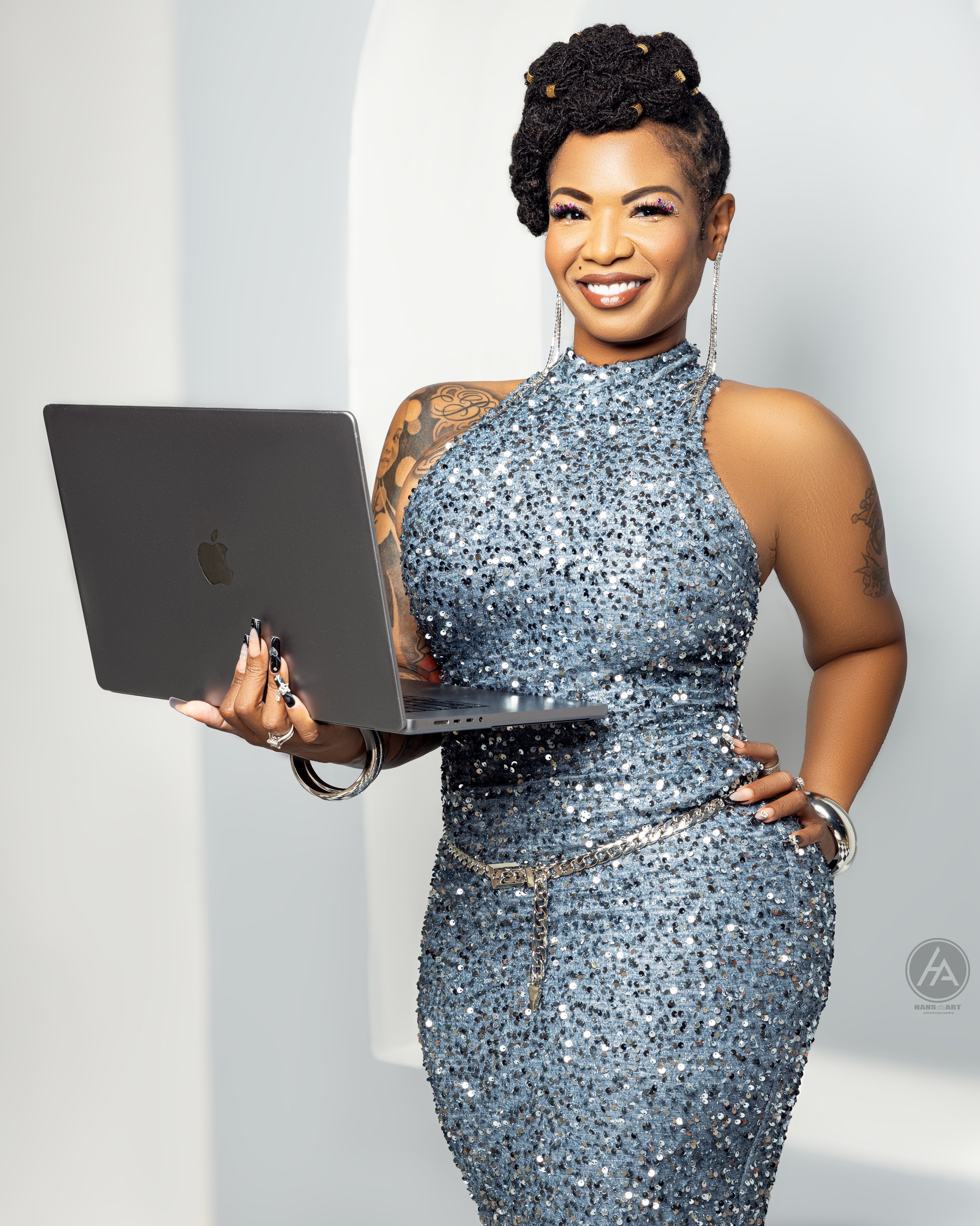 A woman with short, curly hair holding a laptop, wearing a silver sequin dress, smiling, standing against a plain background.