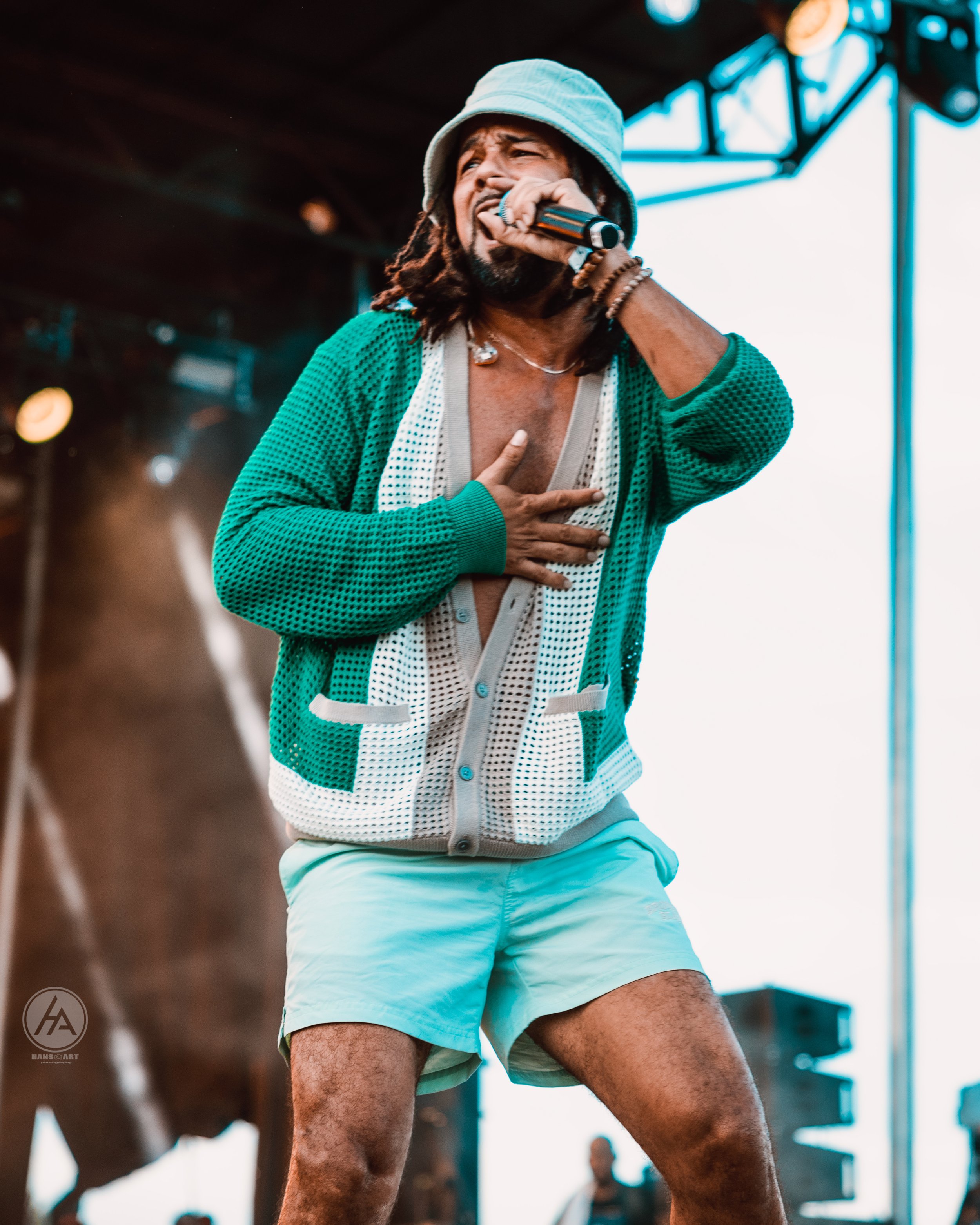 Man with dreadlocks wearing bucket hat, teal cardigan, and shorts, singing passionately into microphone, on stage at outdoor concert.