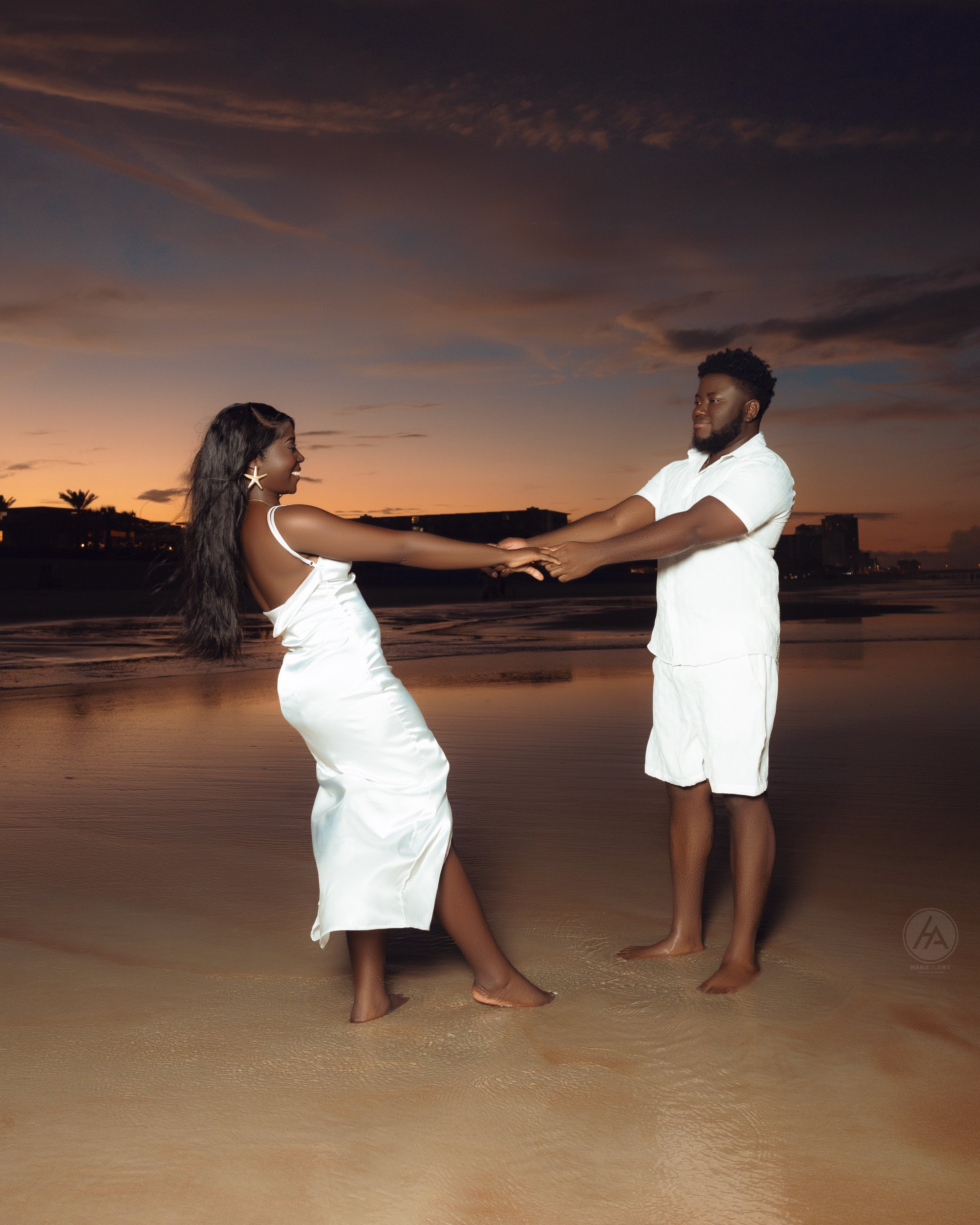 A couple holding hands, dancing on a beach at sunset, with the sky filled with colorful clouds and the water reflecting the sunset.