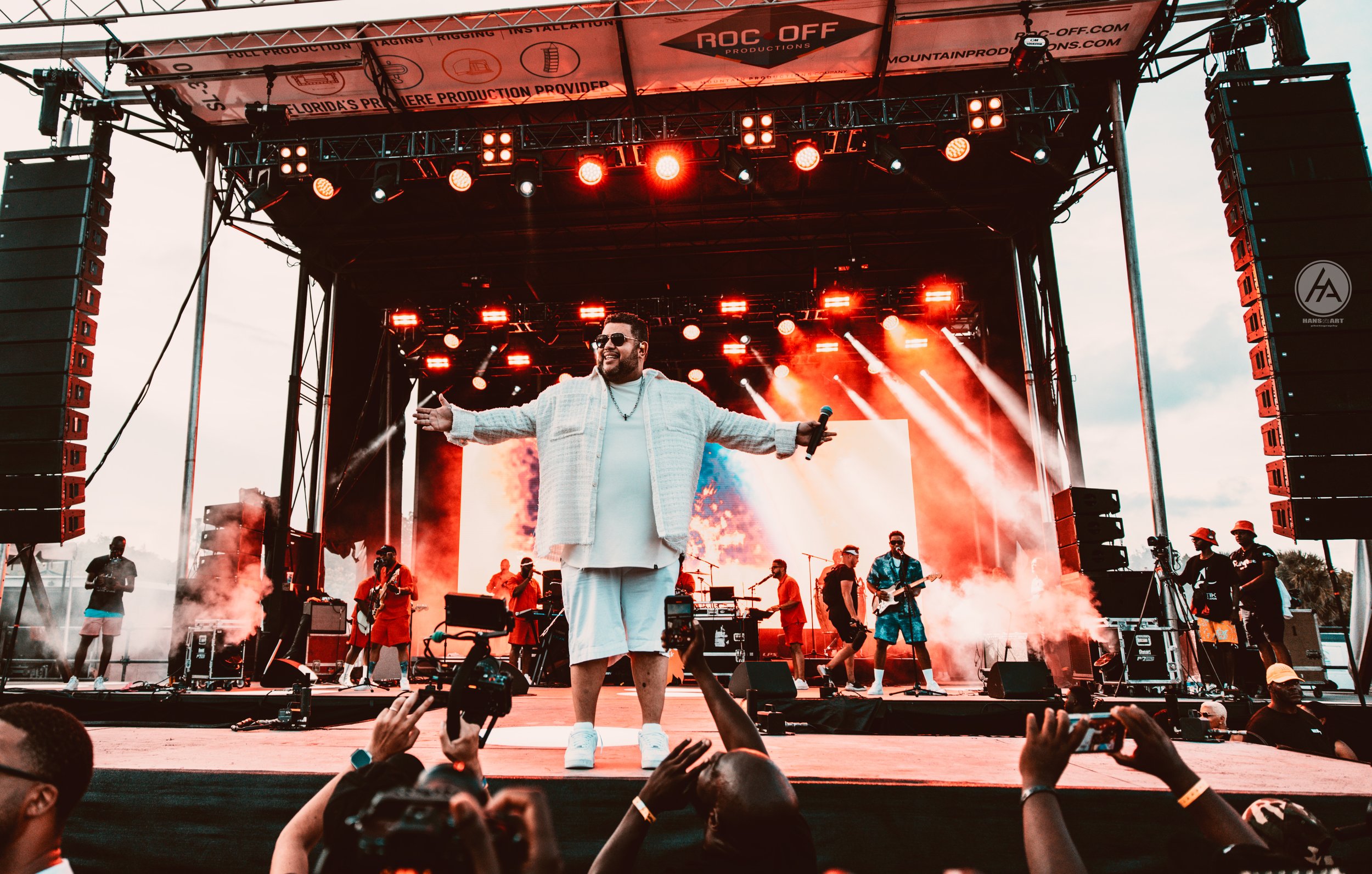 Performer on stage at a music festival with arms outstretched, wearing stylish white outfit, surrounded by musicians and bright red stage lights, crowd taking photos.