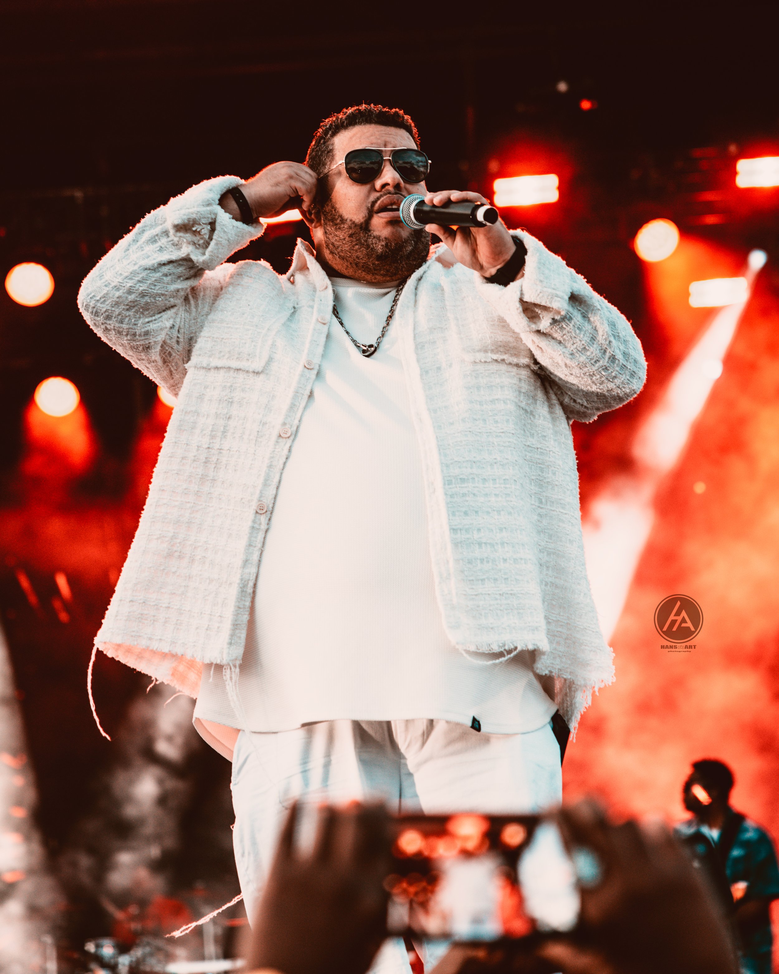 Male performer wearing sunglasses, white textured jacket, white shirt, and a black chain necklace singing into a microphone on stage with red lights in the background.