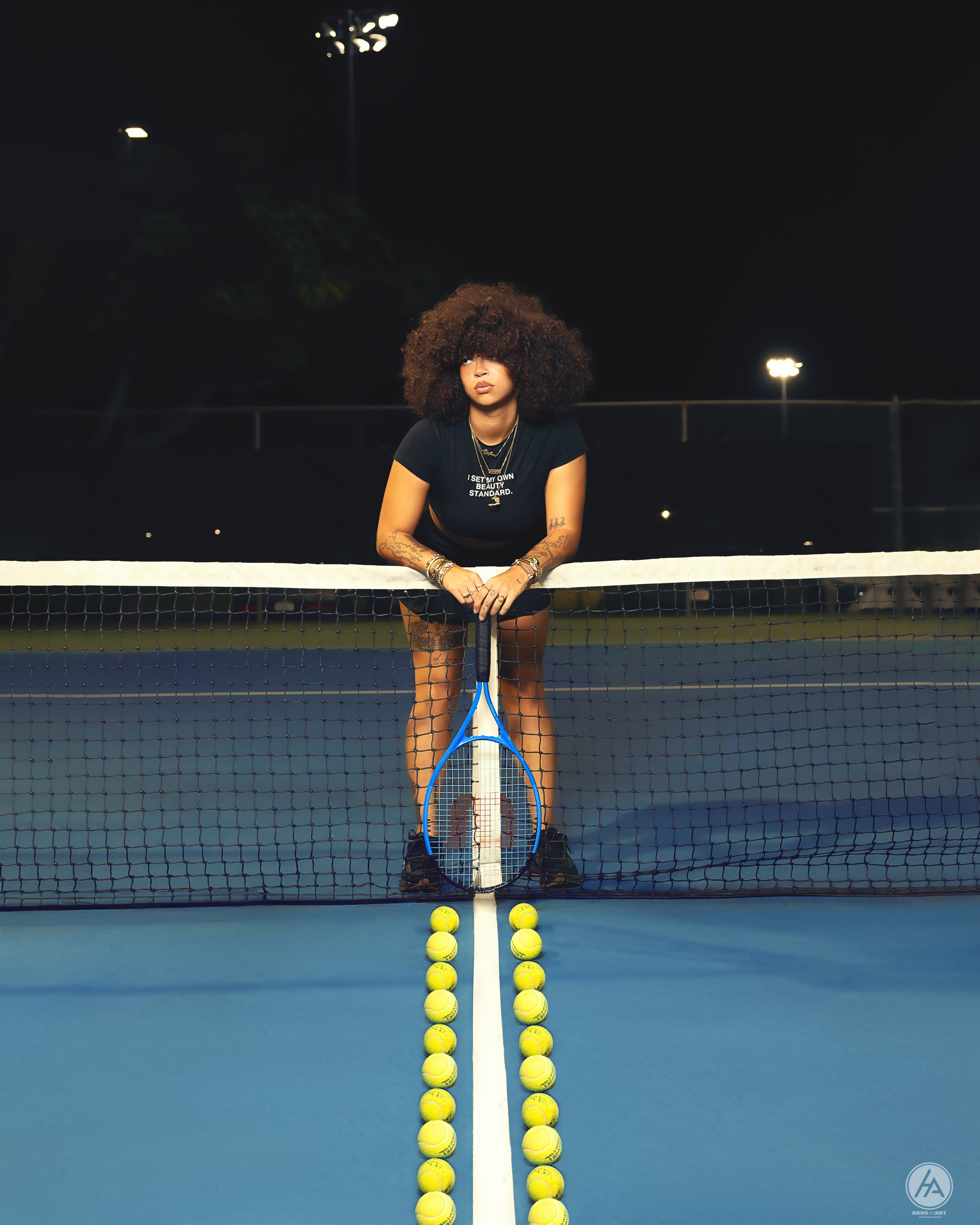 A woman with voluminous curly hair leaning on a tennis net at night, with tennis balls lined up on the court in front of her, holding a tennis racket vertically.