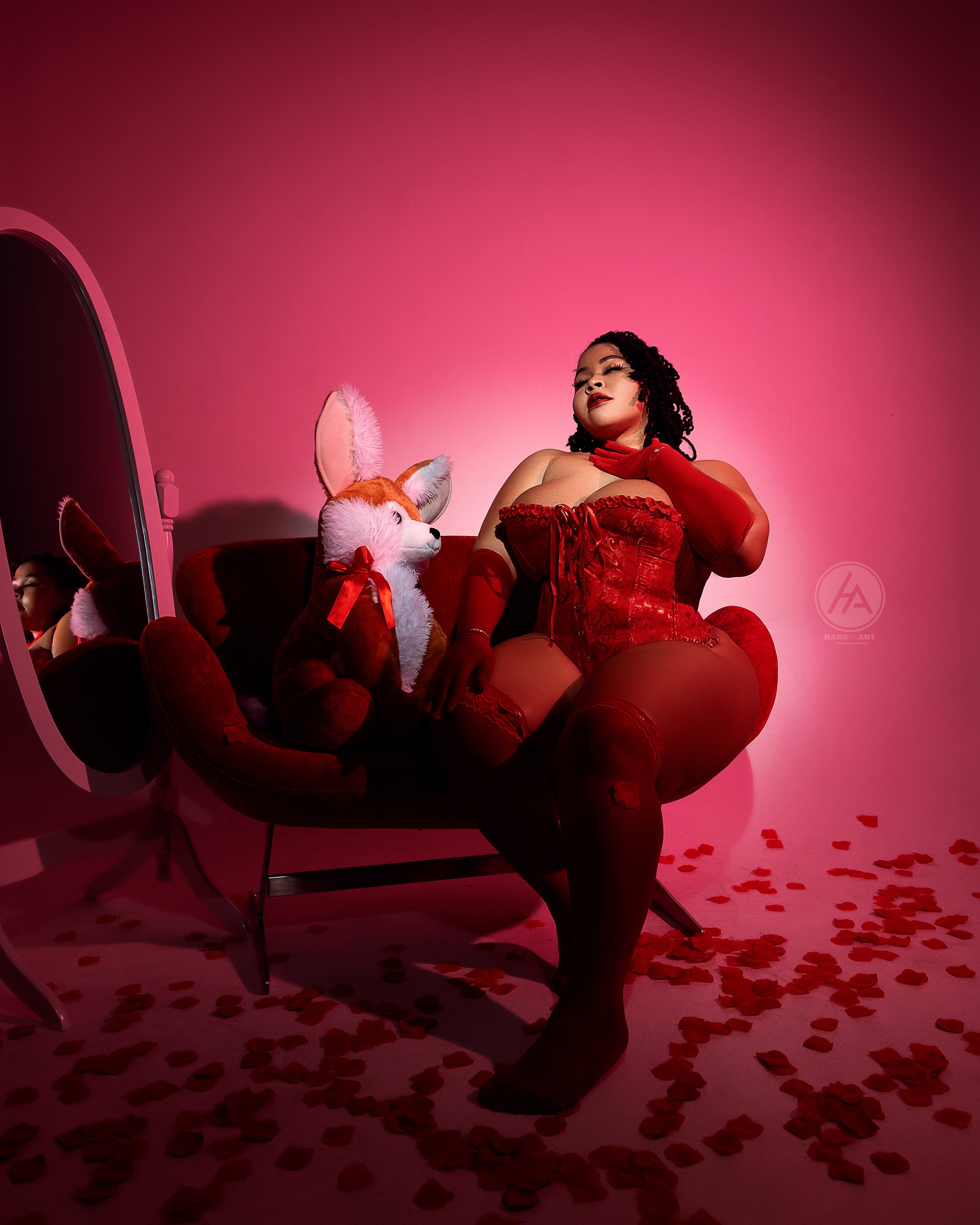 A woman in red lingerie and stockings sitting on a red velvet chair with rose petals on the floor. She is accompanied by a plush fox toy with a red bow, sitting on the chair. There is a large mirror to her left reflecting her face and part of the roo