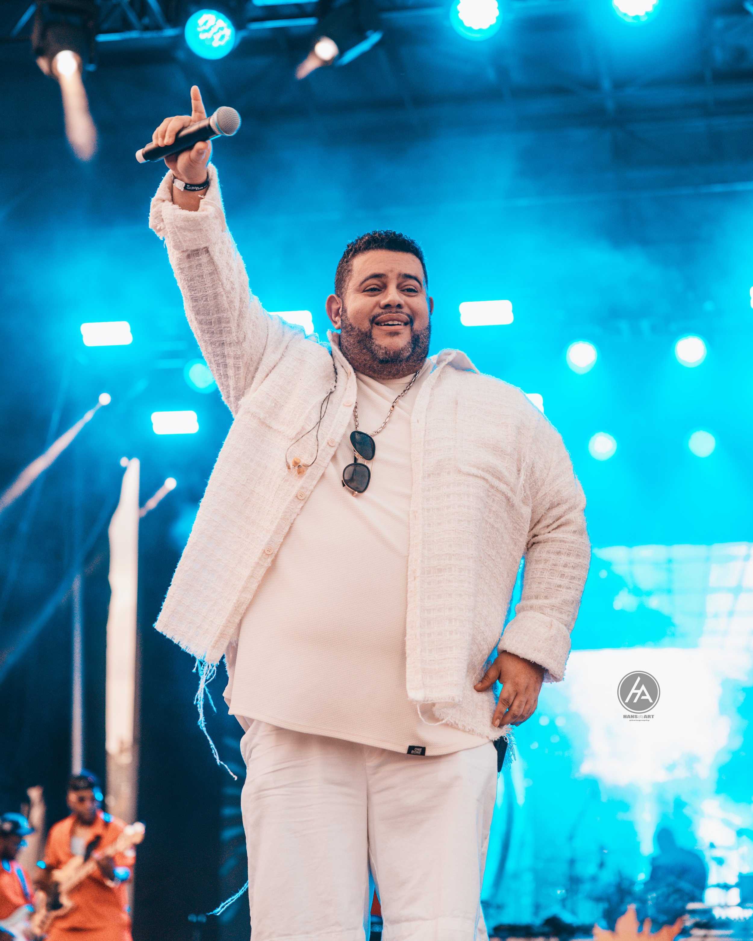 A man in a beige jacket and pants holding a microphone on stage, raising his hand, with stage lights and a band in the background.