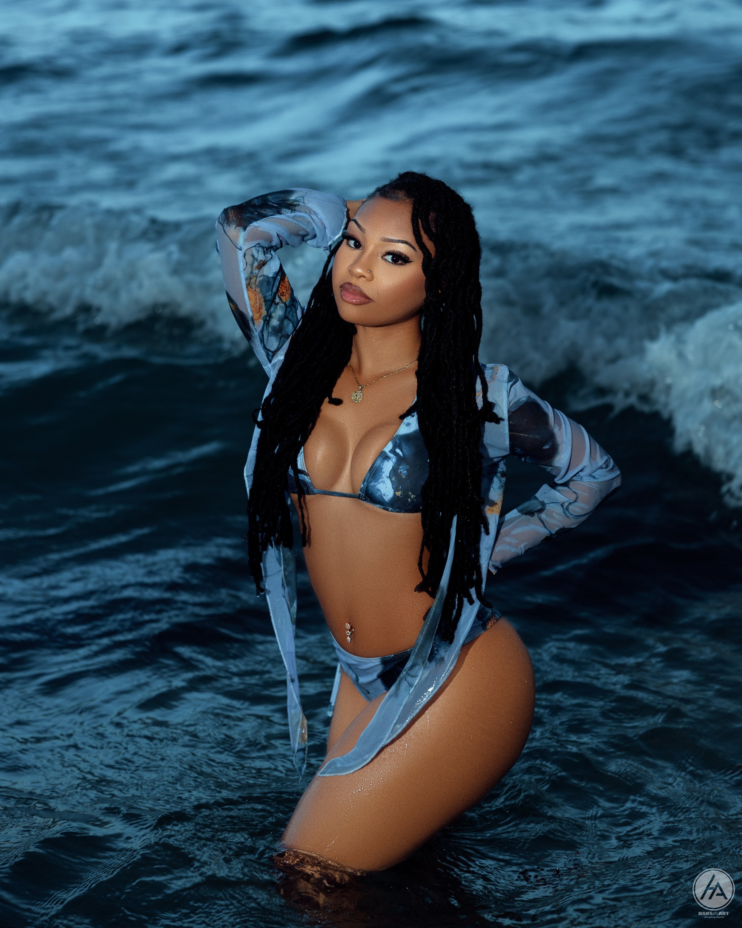 A woman with long black dreadlocks in a blue swimsuit and sheer cover-up kneeling in the ocean at dusk or night, with waves in the background.
