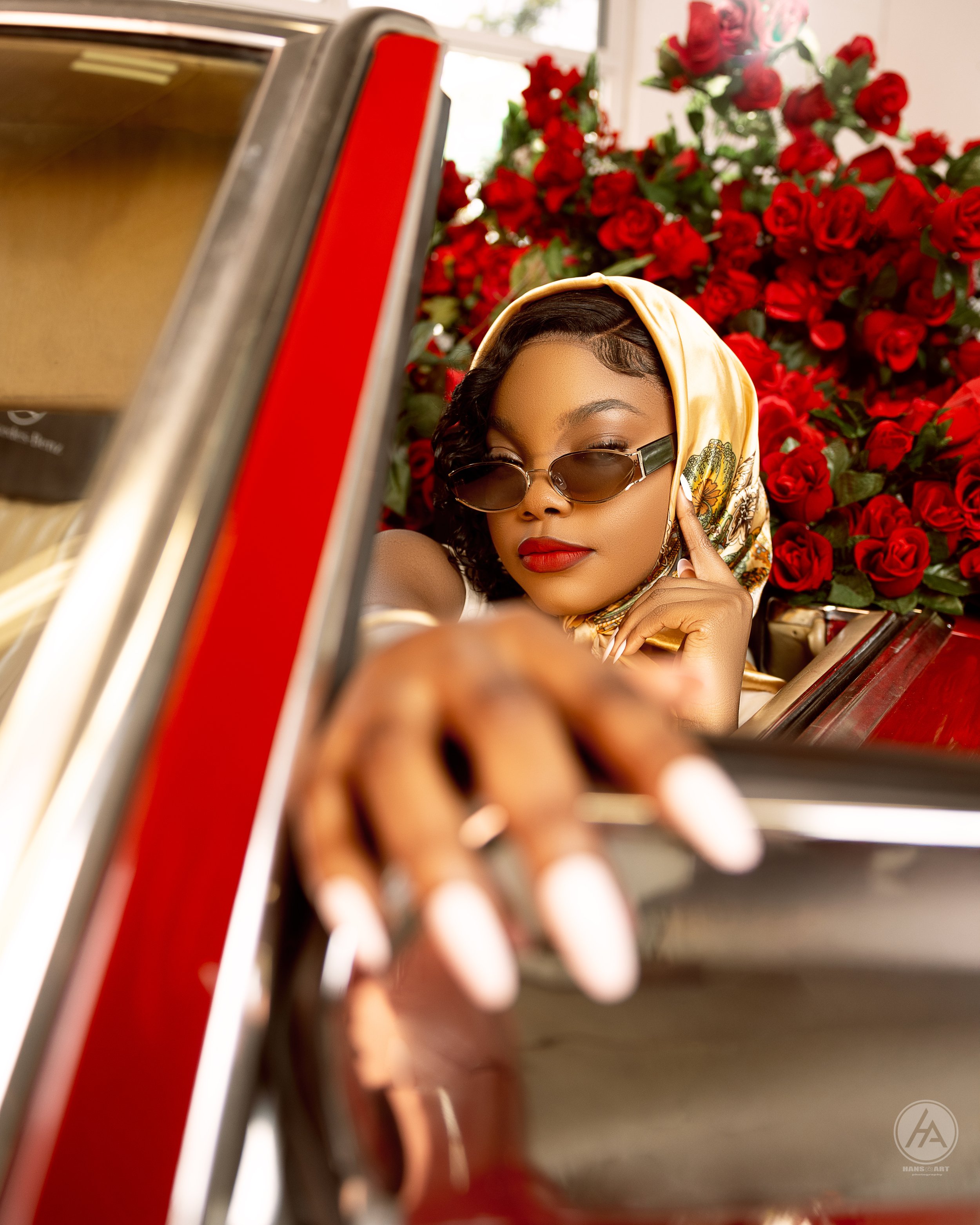 A woman with dark curly hair, wearing sunglasses, a silk headscarf, and red lipstick, takes a selfie with a vintage camera in front of a backdrop of red roses.