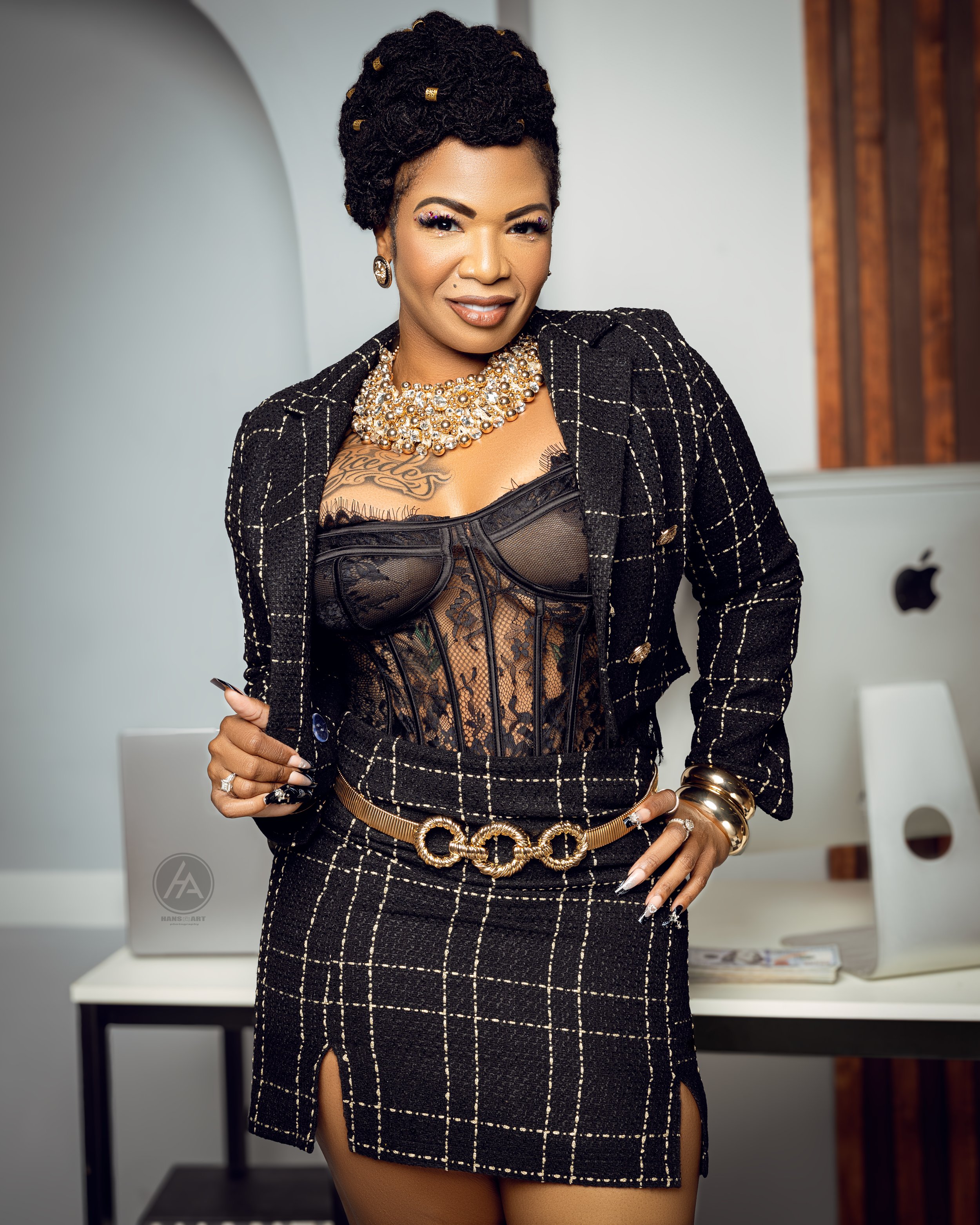 A woman with styled black hair, tattoos, and jewelry, wearing a black lace corset, fitted plaid skirt with high slit, and a black jacket, standing in an office with Apple computers.