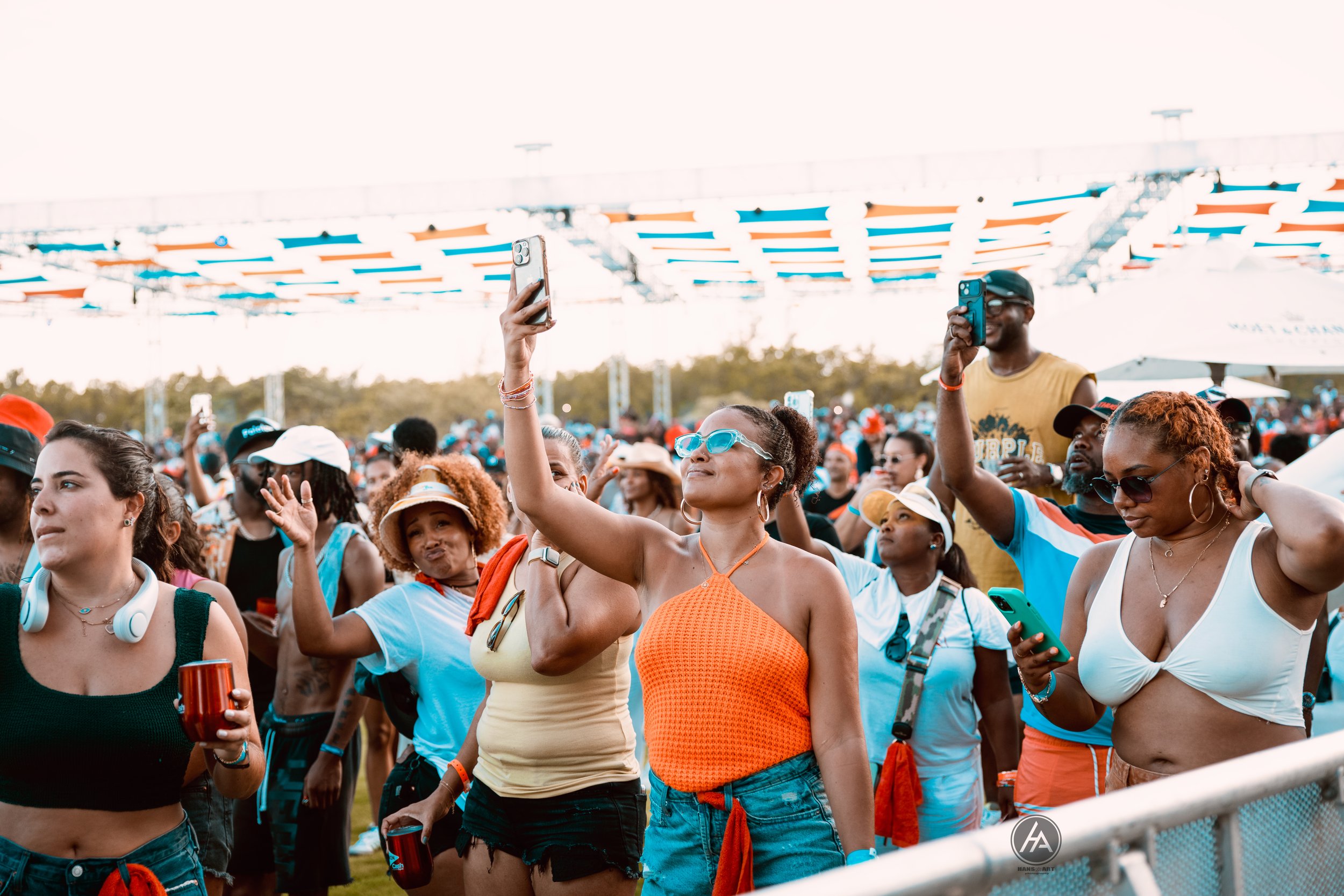 A diverse group of people enjoying an outdoor concert or festival, some taking photos or videos with their smartphones, with a stage or structure in the background under a clear sky.