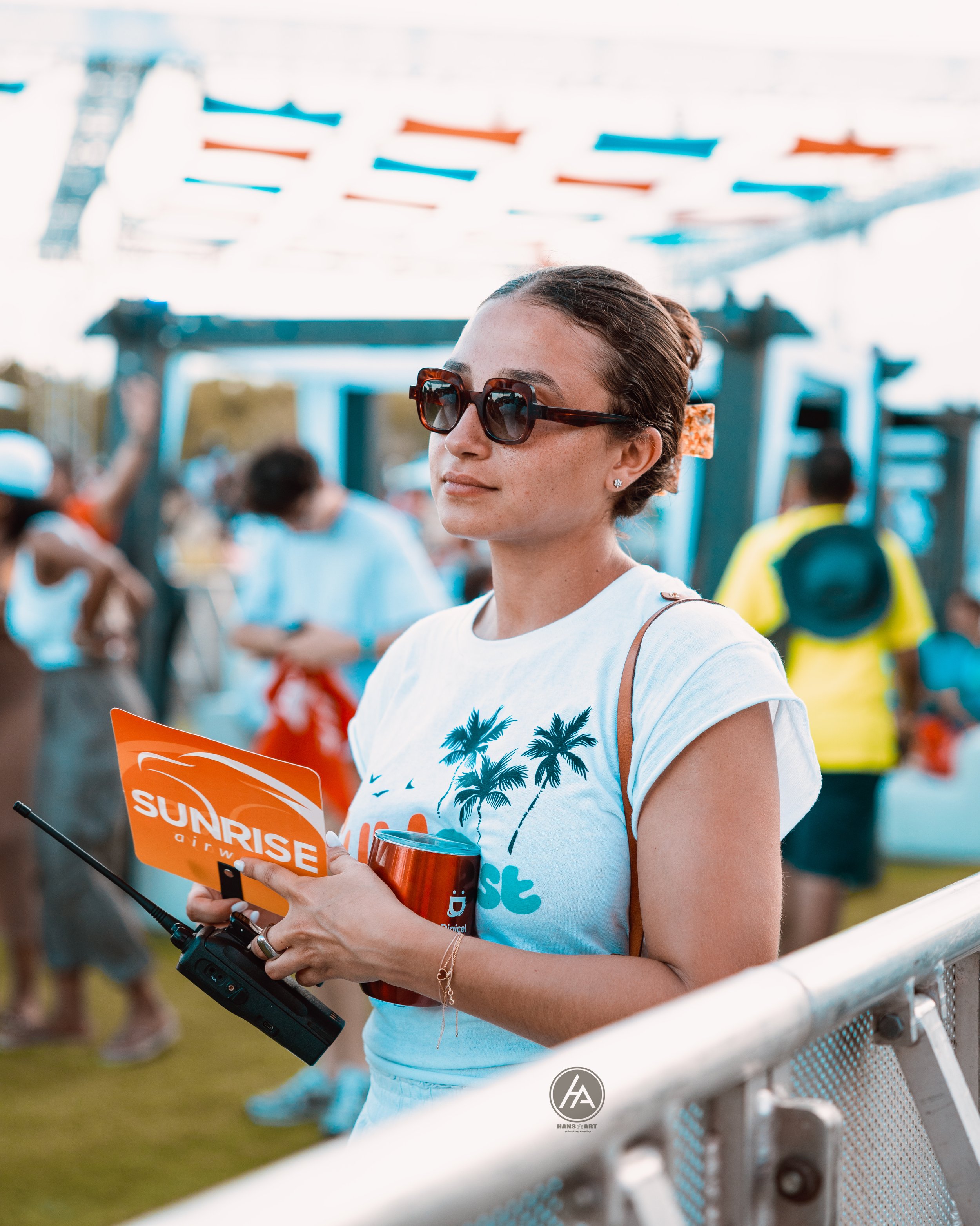 A woman wearing sunglasses and a white t-shirt with palm trees holds a radio and a Sunrise airways pamphlet at an outdoor event, with other people in the background.